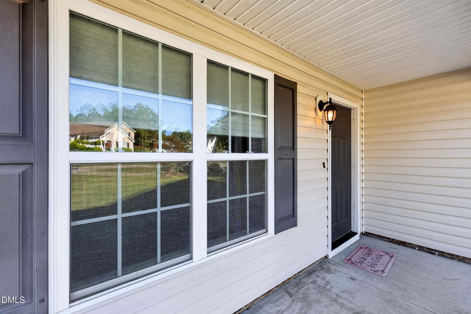 A covered front porch with beige siding, white trim, a large window with multiple panes, a black front door with a lantern-style light, and a welcome mat on concrete flooring.