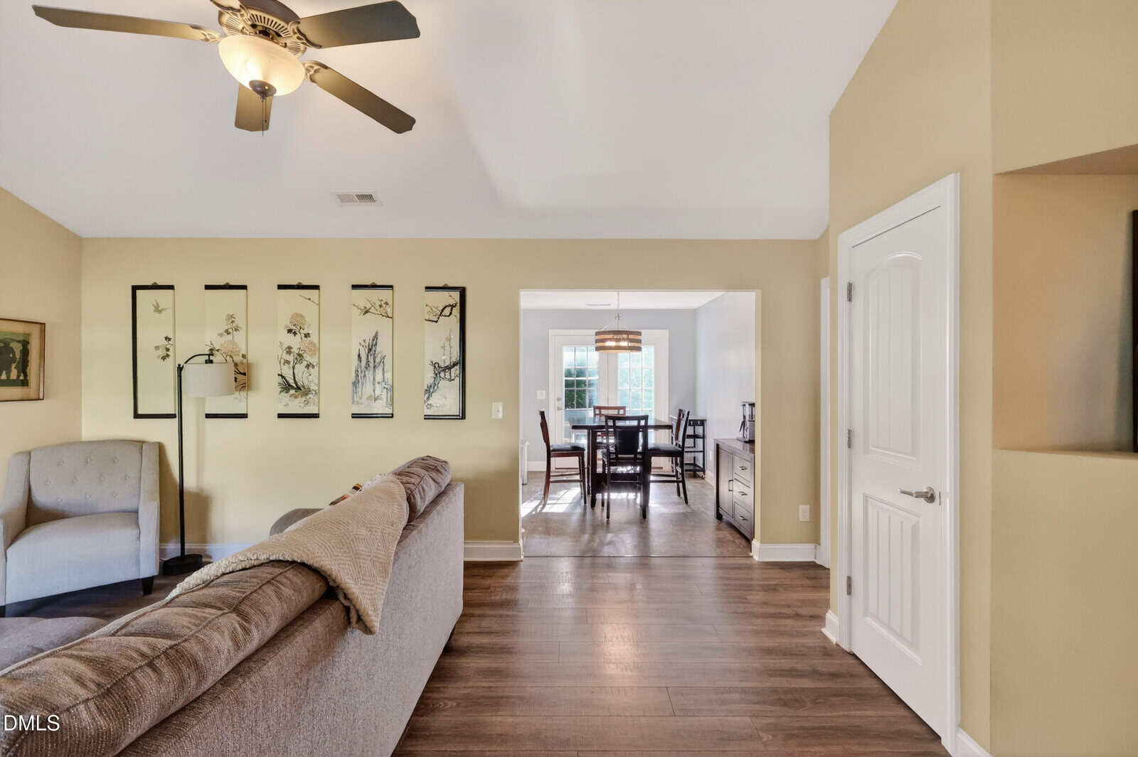 A cozy living room with a ceiling fan, sectional sofa, armchair, and wall art opens to a dining area with a large window, wooden table, and modern chandelier. The space features wood floors and neutral-colored walls.