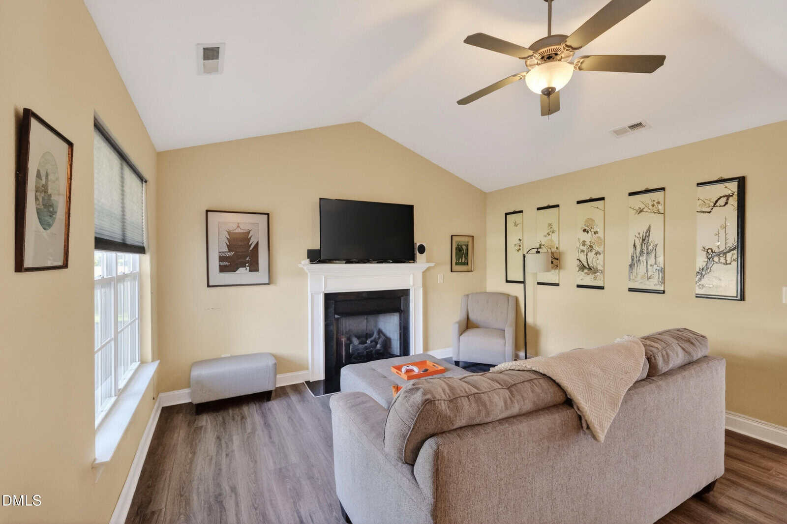 A cozy living room with a gray sofa, armchair, fireplace, wall-mounted TV, and Asian-inspired artwork on beige walls. A ceiling fan hangs from the angled ceiling above wood flooring.