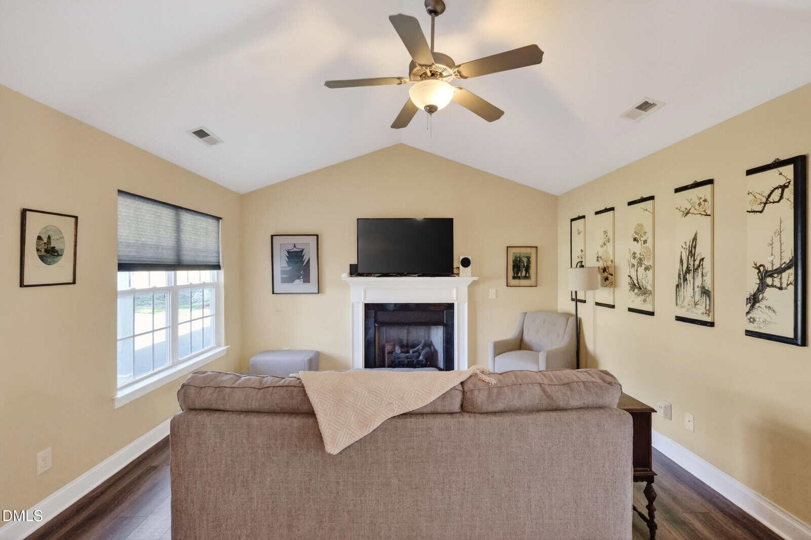 A cozy living room with a sofa, armchair, and ottoman facing a fireplace and TV. Art hangs on the tan walls, and a window with blinds lets in natural light. A ceiling fan is above.
