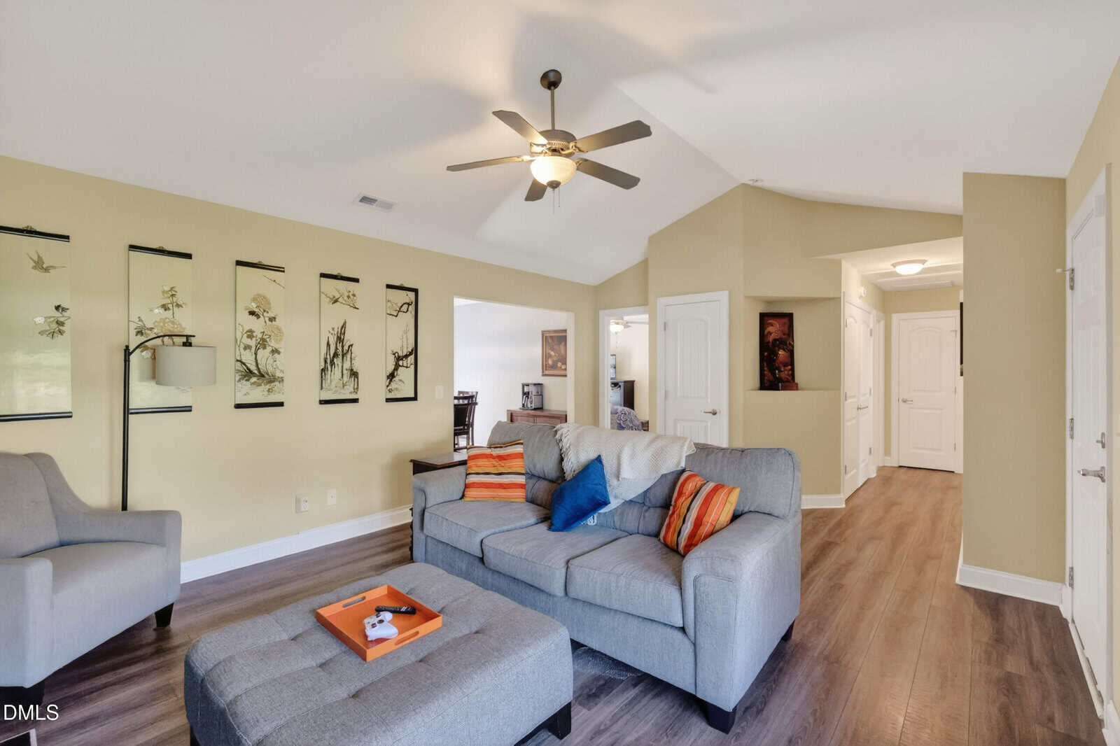A cozy living room with gray sofas, colorful pillows, a ceiling fan, wall art, and wood flooring. An open doorway leads to a dining area, and a hallway with white doors is visible in the background.