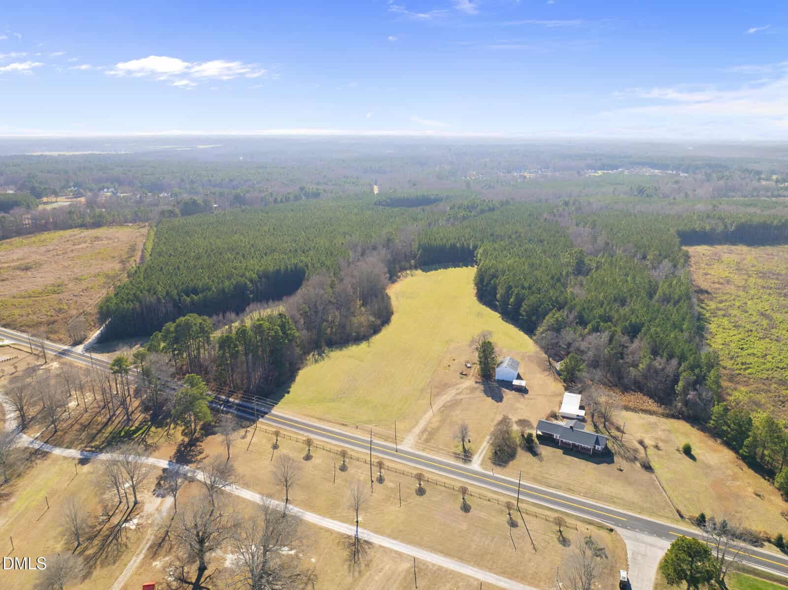 Aerial view of a rural landscape featuring a large open field surrounded by dense trees, a few houses and buildings, and a road running across the foreground under a blue sky with scattered clouds.