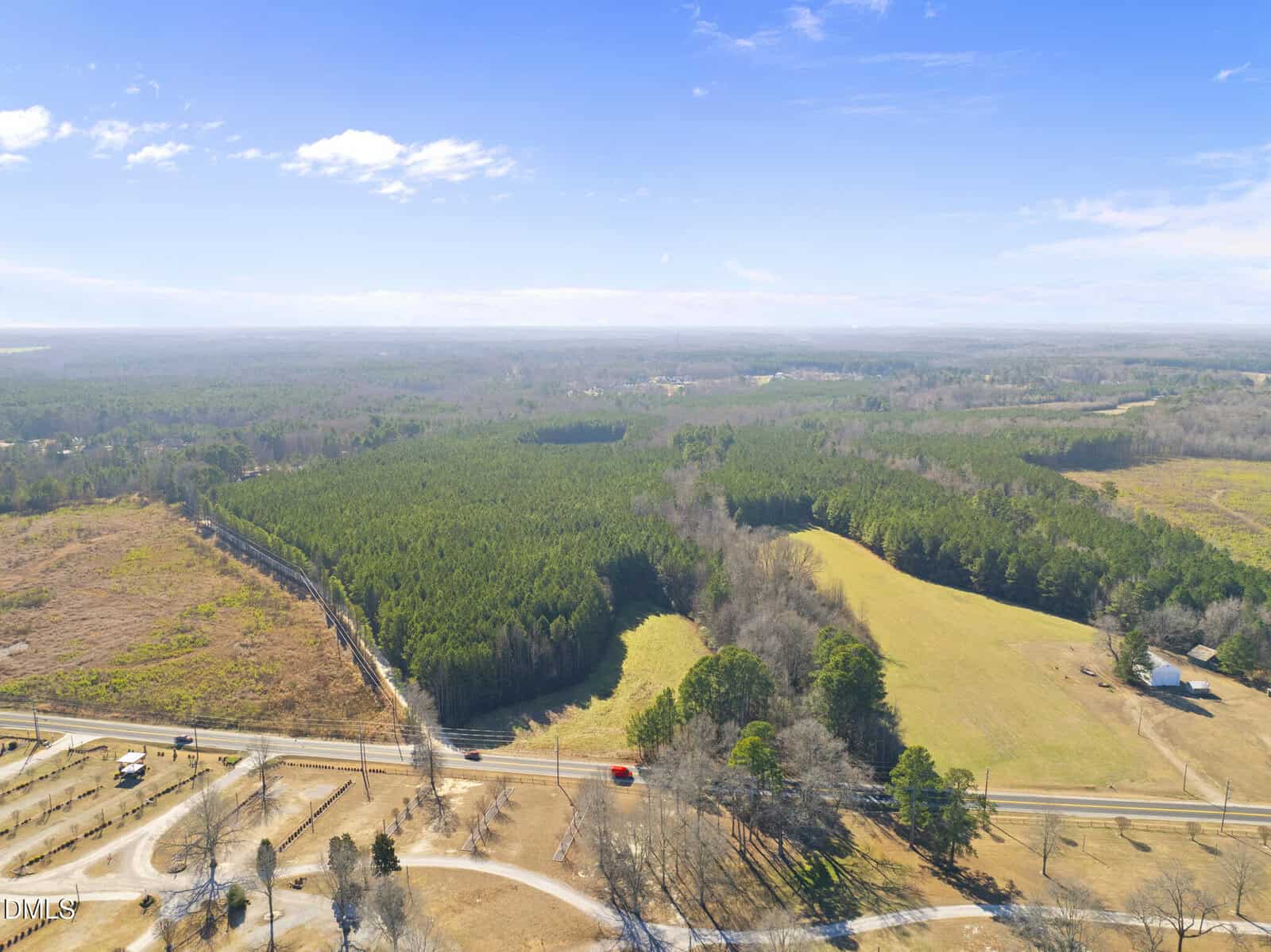 Aerial view of a rural landscape with a mix of dense green forest, open grassy fields, a few scattered trees, a country road, and a small structure near the road under a clear blue sky.