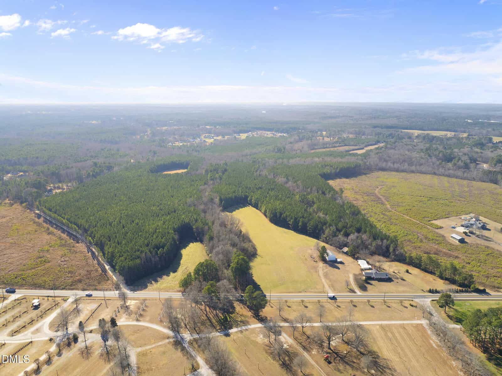 Aerial view of a rural landscape with open fields, patches of forest, a few houses, and a road running through the foreground under a bright blue sky.