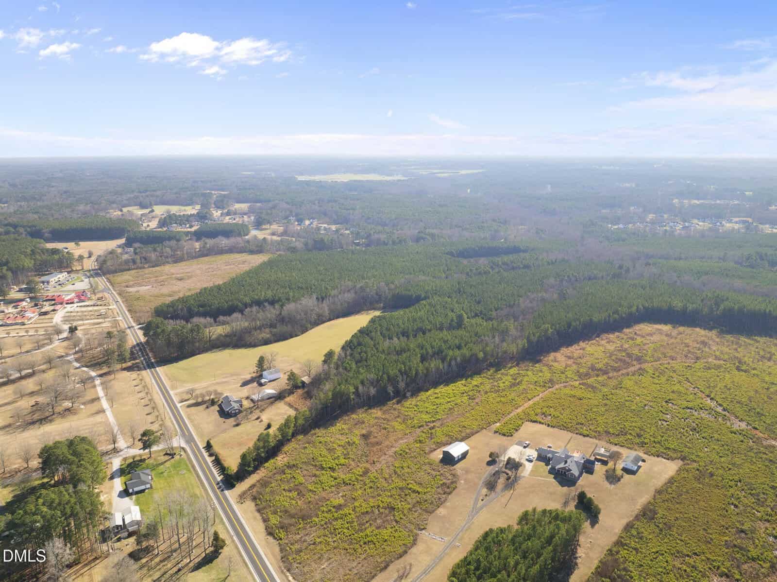 Aerial view of a rural landscape with a road, scattered houses, fields, and wooded areas under a partly cloudy sky, stretching toward the horizon.