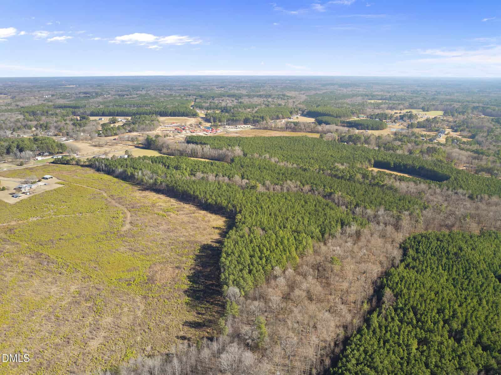 Aerial view of a rural landscape with forests, open fields, and scattered buildings under a clear blue sky. Dense green trees and leafless patches are visible, with distant structures near the horizon.