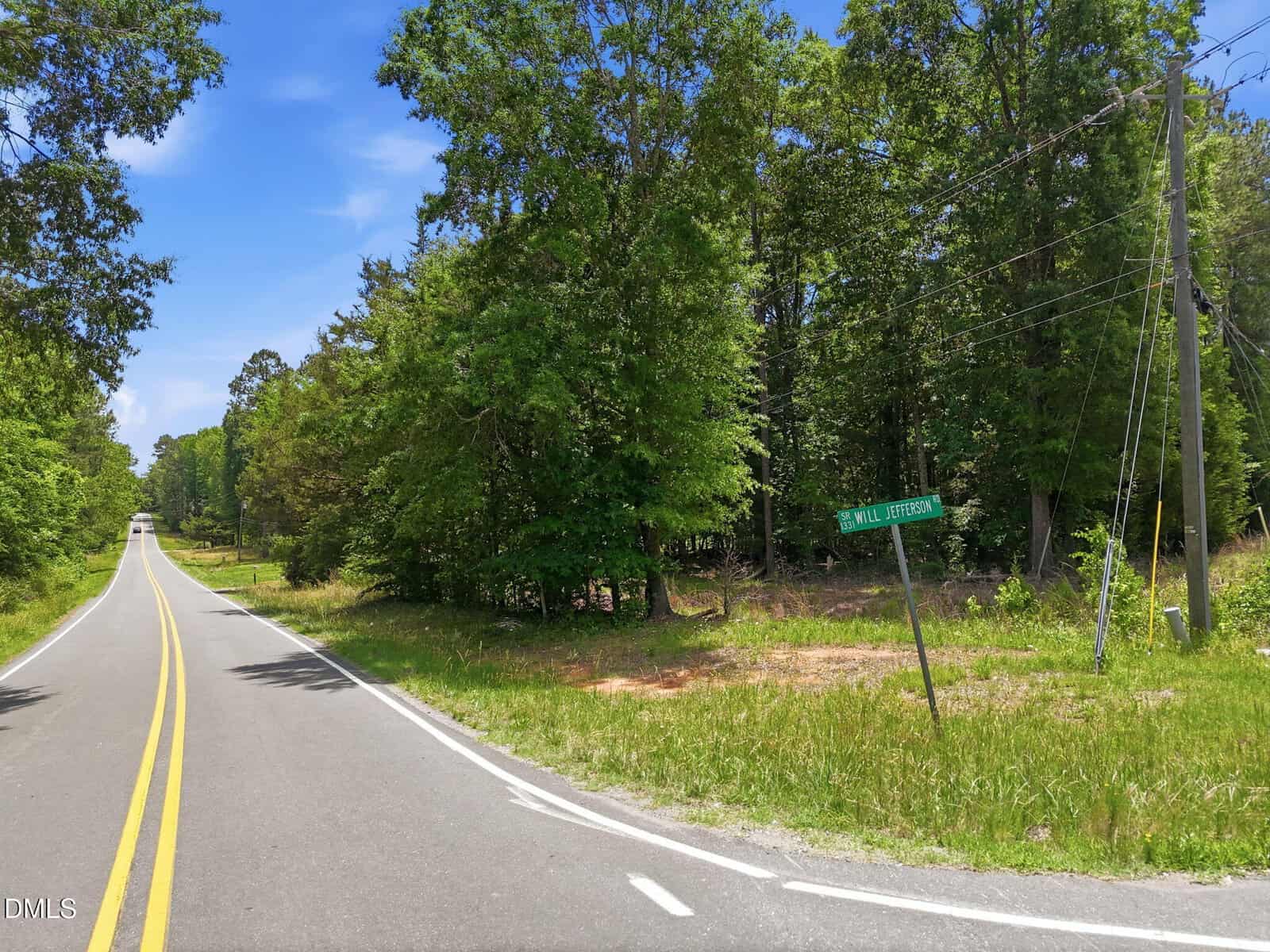 A two-lane rural road curves past tall green trees under a blue sky. A green street sign reading "WILL JEFFERSON RD" is visible on the right near the edge of a grassy area with power lines.