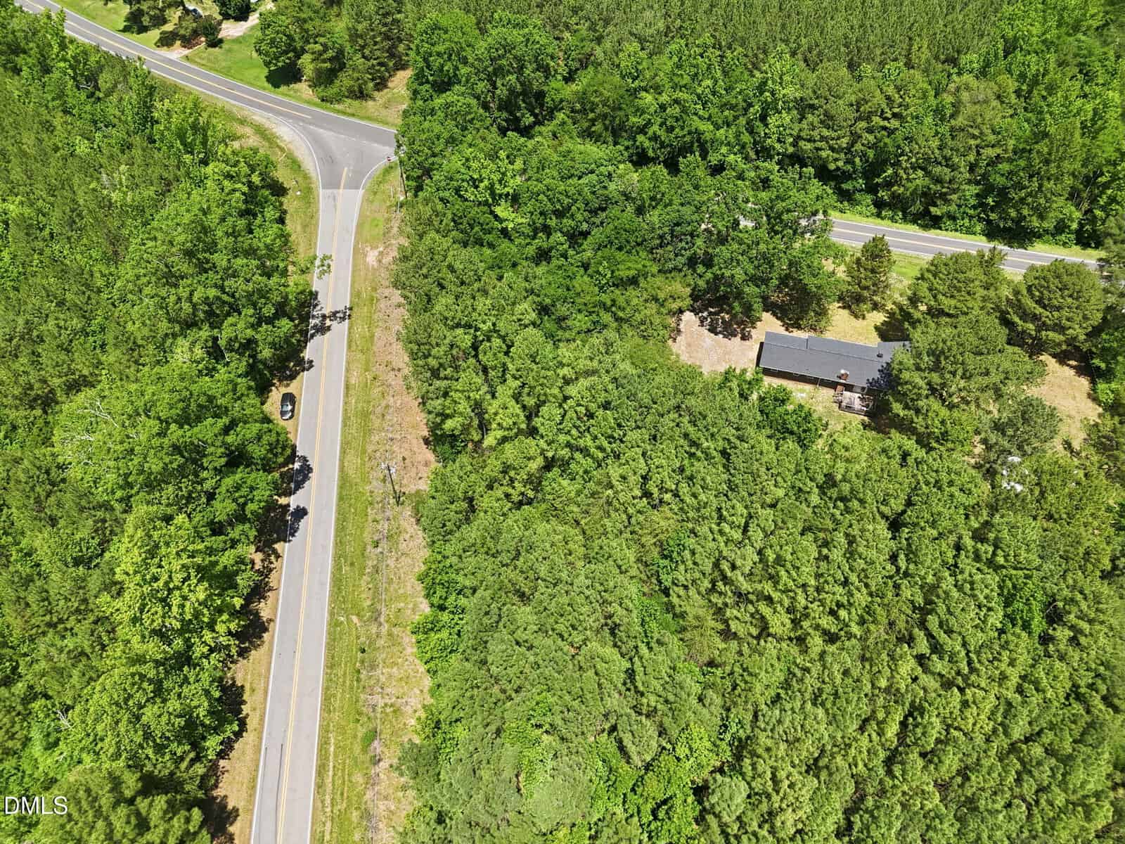 Aerial view of a wooded area with two intersecting roads, one car on the left road, and a house partially visible among the trees on the right side.