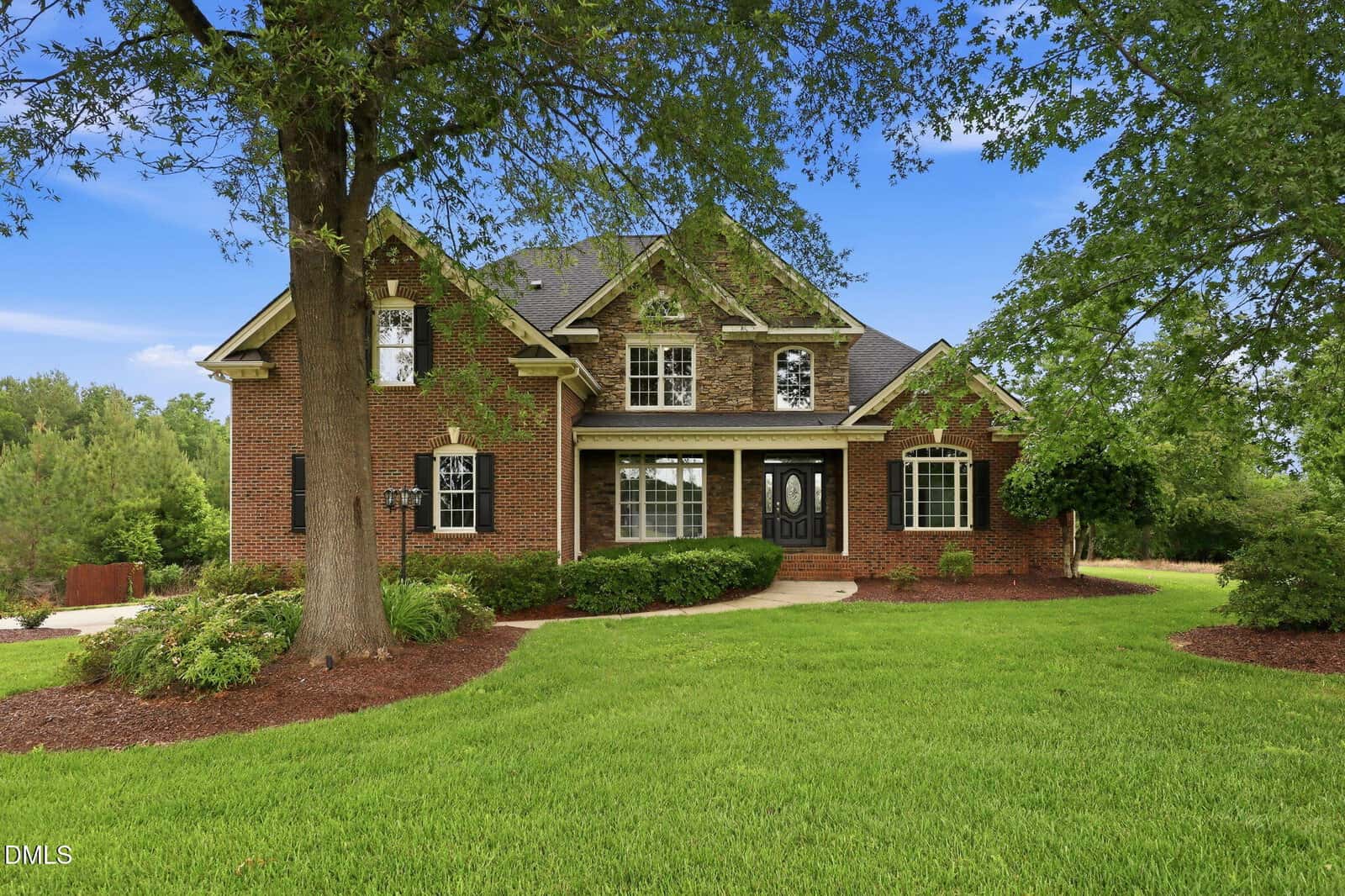 Two-story red brick house with black shutters, large windows, and a covered front porch. The home sits on a well-maintained lawn with trees, shrubs, and a curved walkway leading to the entrance.