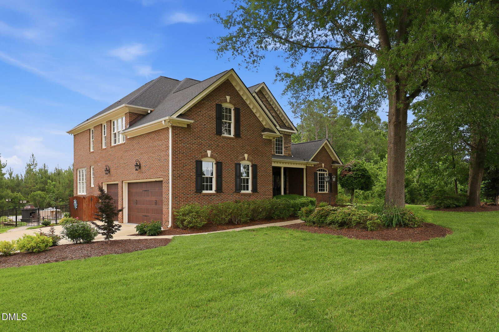 A two-story brick house with black shutters and a two-car garage sits on a well-maintained lawn with mulch, shrubs, and large trees under a blue sky.