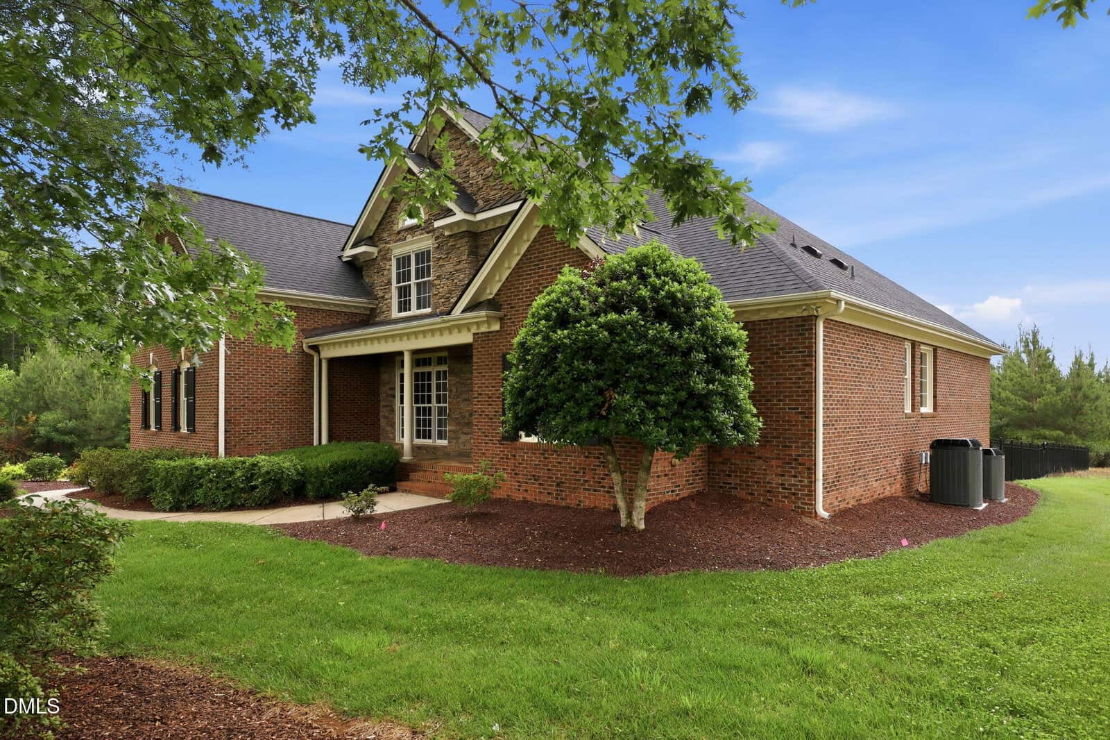A brick house with a manicured lawn, shrubs, and a small tree in front, under a blue sky with scattered clouds. The home has multiple rooflines and a covered front entryway.