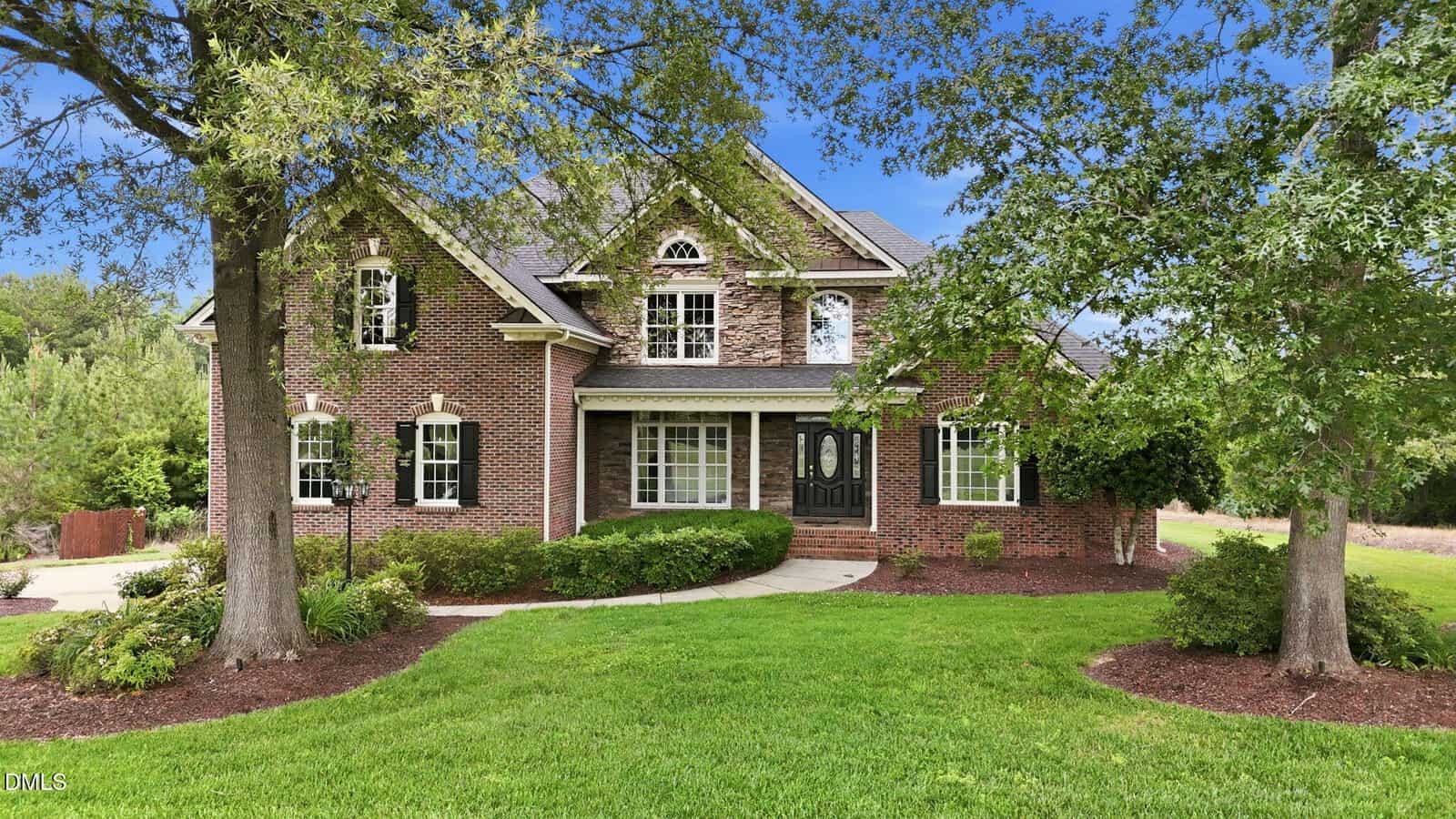 A two-story brick house at 1257 Pine Ridge Road in Zebulon, with black shutters and a black front door, sits among green grass, shrubs, and tall trees beneath a blue sky. A curved walkway leads to the inviting entrance.