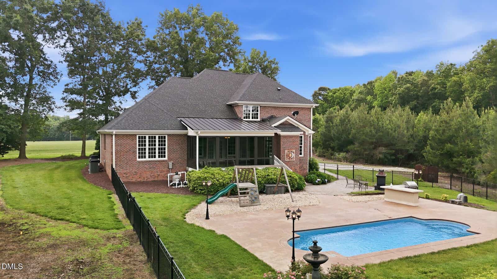 A two-story brick house with a fenced backyard features a patio, in-ground swimming pool, grill area, and a small slide. The yard is bordered by trees and a lawn under a clear blue sky.
