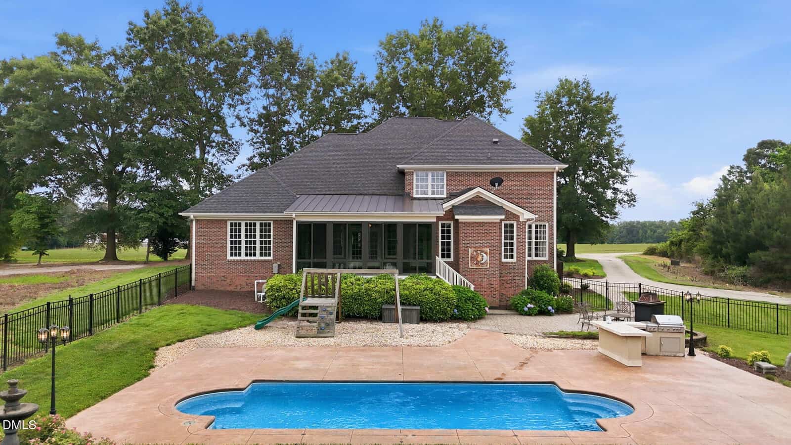 A brick house with a screened porch, fenced backyard, outdoor pool, built-in grill, and patio area, surrounded by trees and a lawn under a clear sky.
