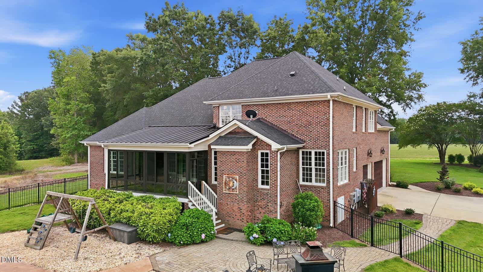 A large two-story brick house with a screened porch, fenced backyard, swing set, and landscaped garden, set against trees and a spacious green lawn under a blue sky.