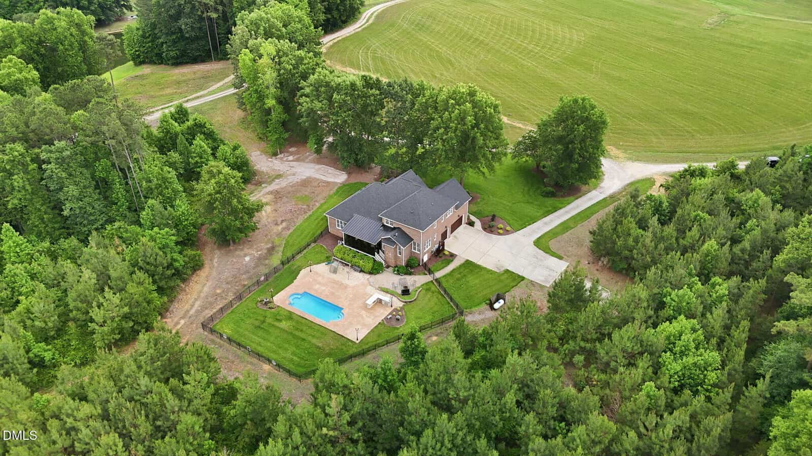 Aerial view of a large brick house with a driveway, surrounded by trees and fields. A fenced backyard with a swimming pool and patio is visible.