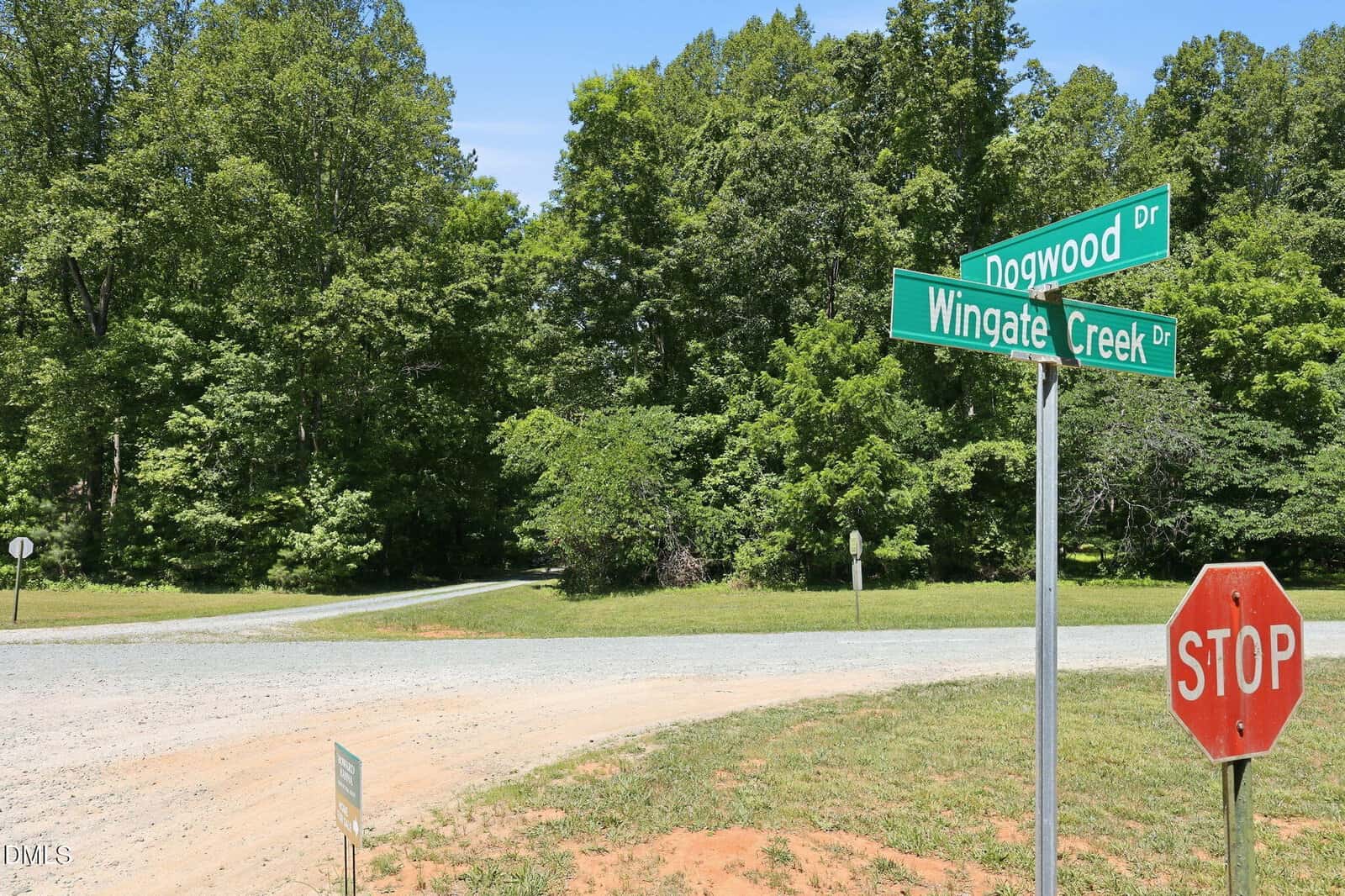 A stop sign stands at the corner of Dogwood Drive and Wingate Creek Drive, with gravel roads and dense green trees in the background under a clear blue sky.