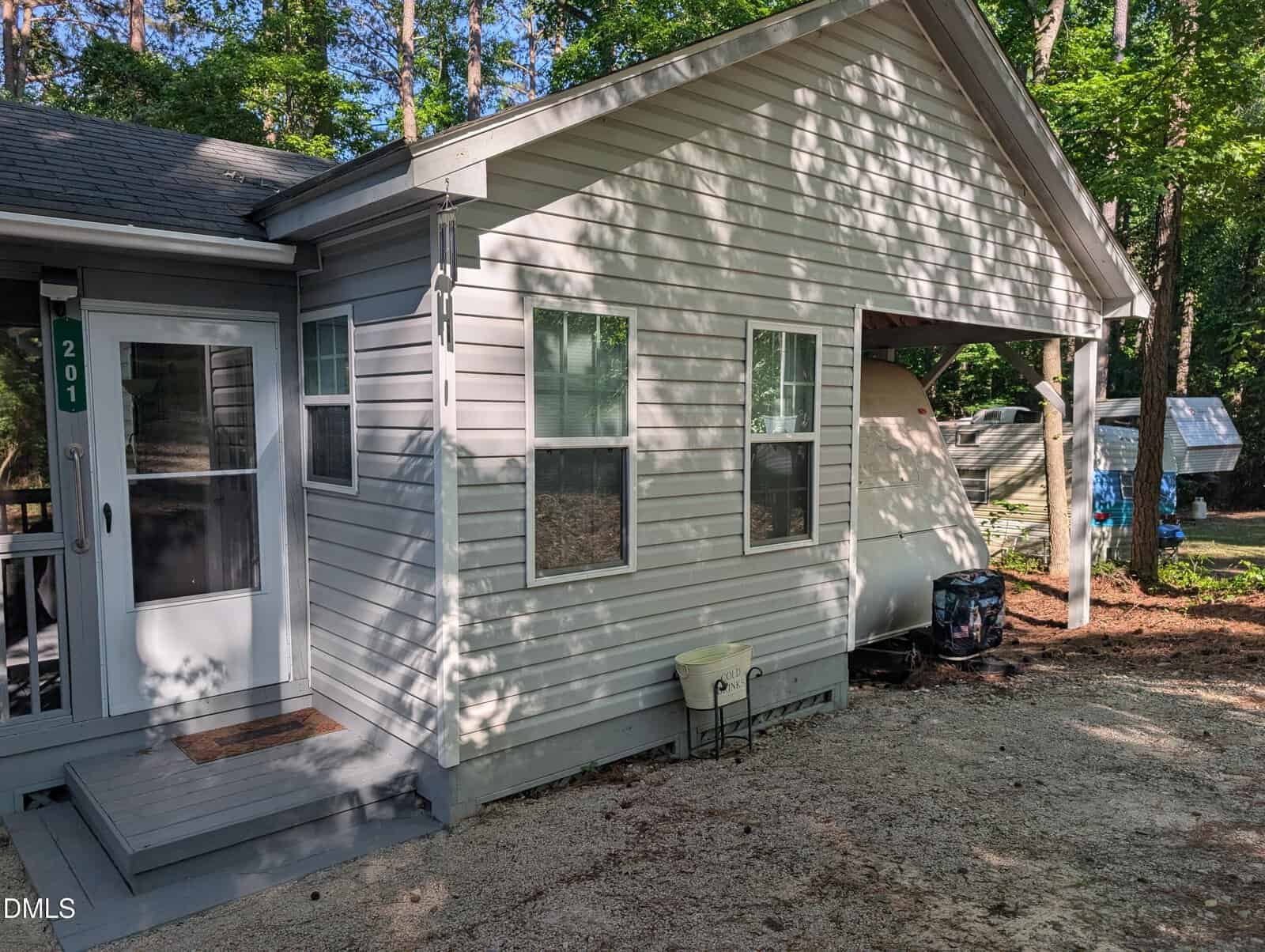 A small gray house with white trim and a covered carport sits among tall trees. The carport shelters a white camper and various outdoor items. Sunlight and shadows are visible on the house and ground.