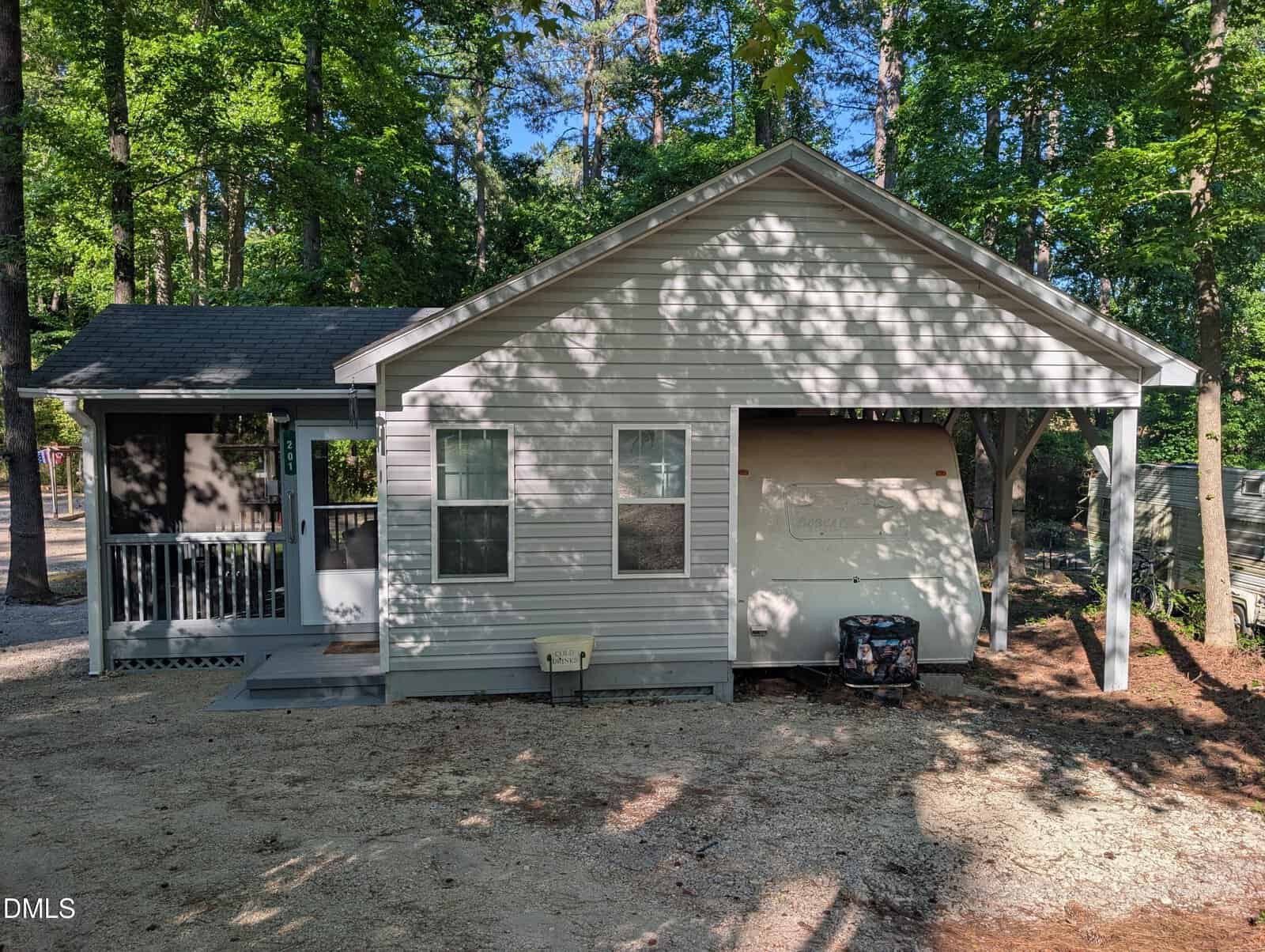 A small single-story gray house with a covered porch sits at 201 Running Deer Drive, Louisburg, shaded by tall trees. The yard is mostly dirt with patches of sunlight, and an air conditioning unit rests beside the house.