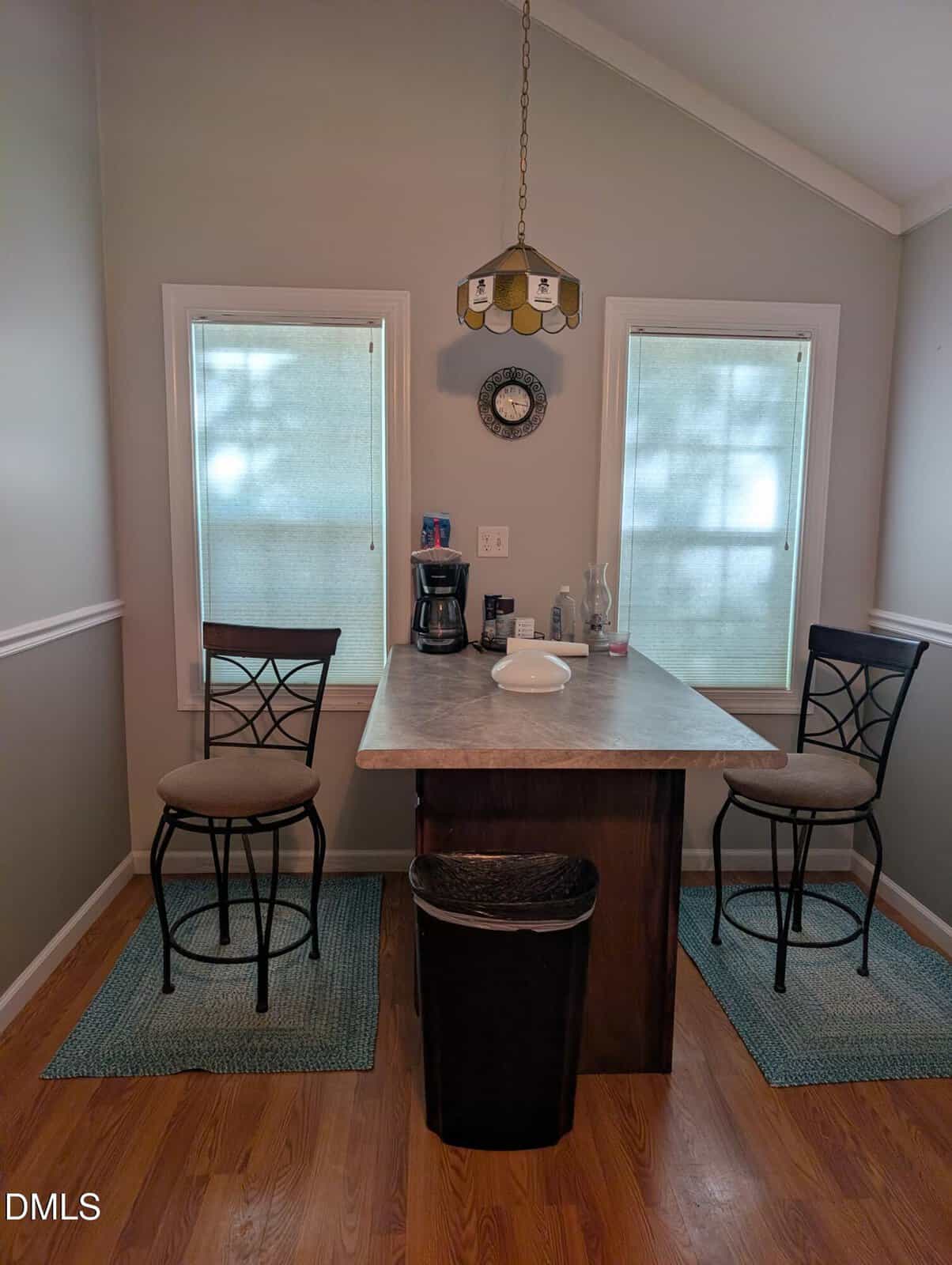 A small dining area with two high chairs at a rectangular table, a coffee maker, bottles, and a plate on the tabletop, set between two windows with blinds. A clock and hanging light fixture are on the wall above.
