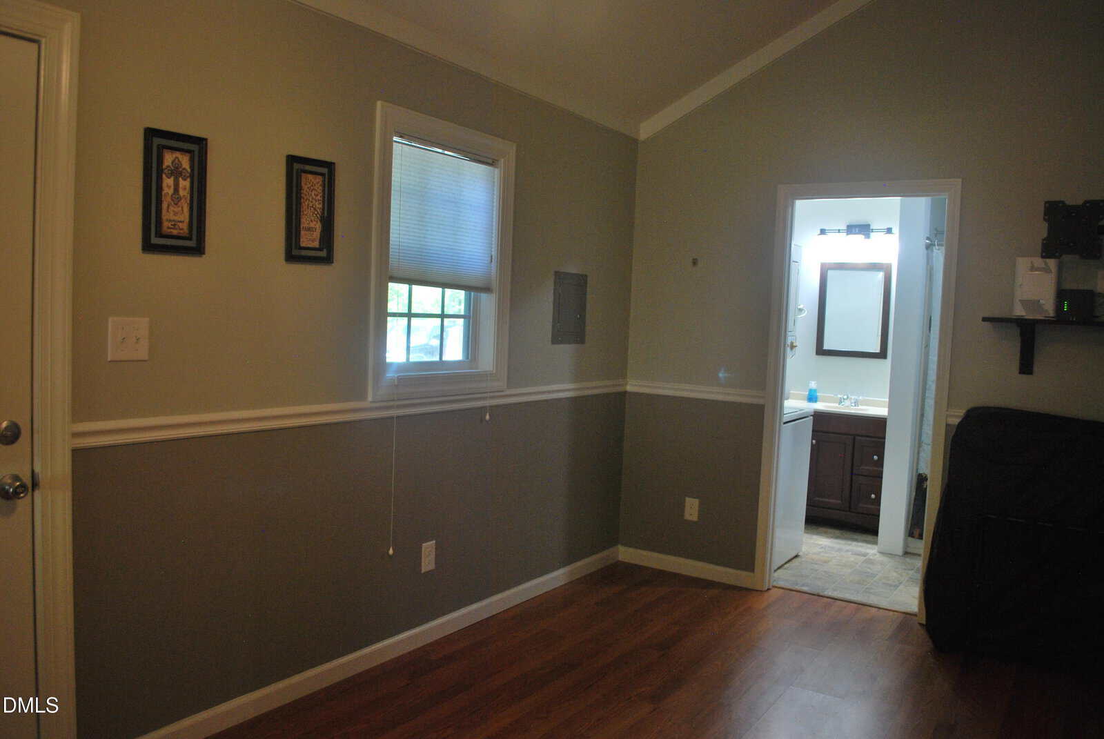 A room with gray walls, wood flooring, a window with blinds, two framed pictures, and an open doorway leading to a bathroom with a sink, mirror, and tiled floor. A shelf and folded item are visible on the right.