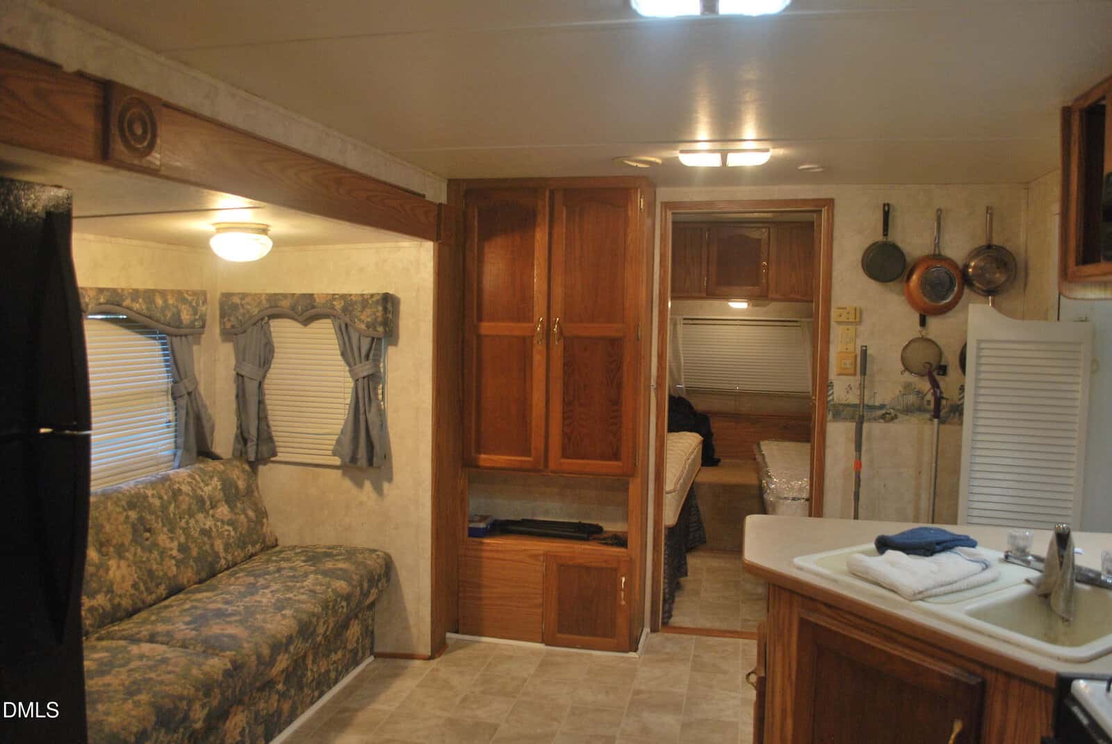 Interior of an RV featuring a floral sofa, wood cabinets, a small kitchen area with hanging pots and pans, and a back room with a bed partly visible. Warm lighting and window treatments add a cozy feel.