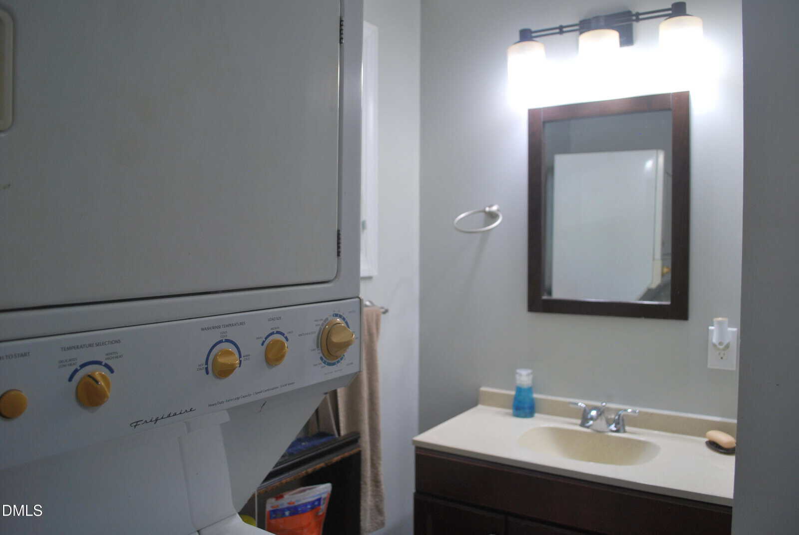 A bathroom with a sink, mirror, towel rack, and a stacked washer and dryer unit visible on the left side. The countertop has soap and a dispenser next to the faucet.
