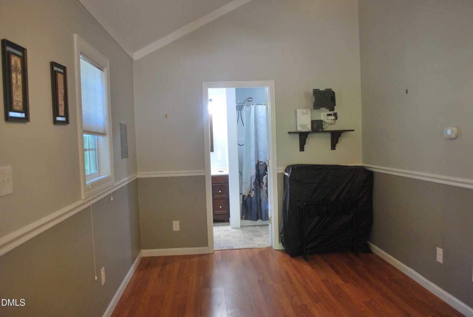 A small room with gray walls, wood flooring, two windows, and a door leading to a bathroom. A folded black futon or mattress leans against the wall under a shelf, and framed art hangs by the windows.