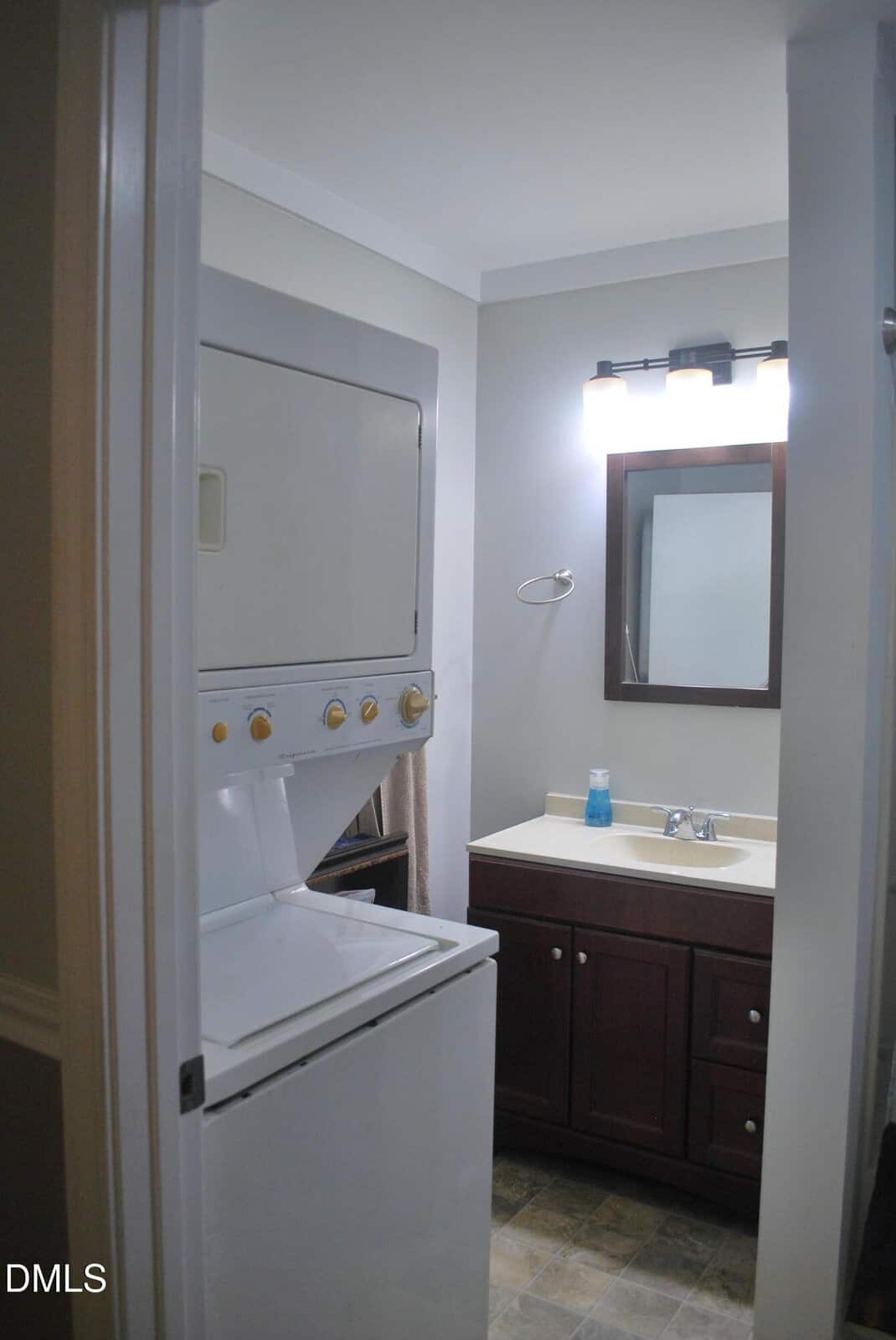 A small bathroom with a stacked washer and dryer next to a sink, mirror, and dark wood vanity. The floor is tiled, and a soap dispenser sits on the counter.