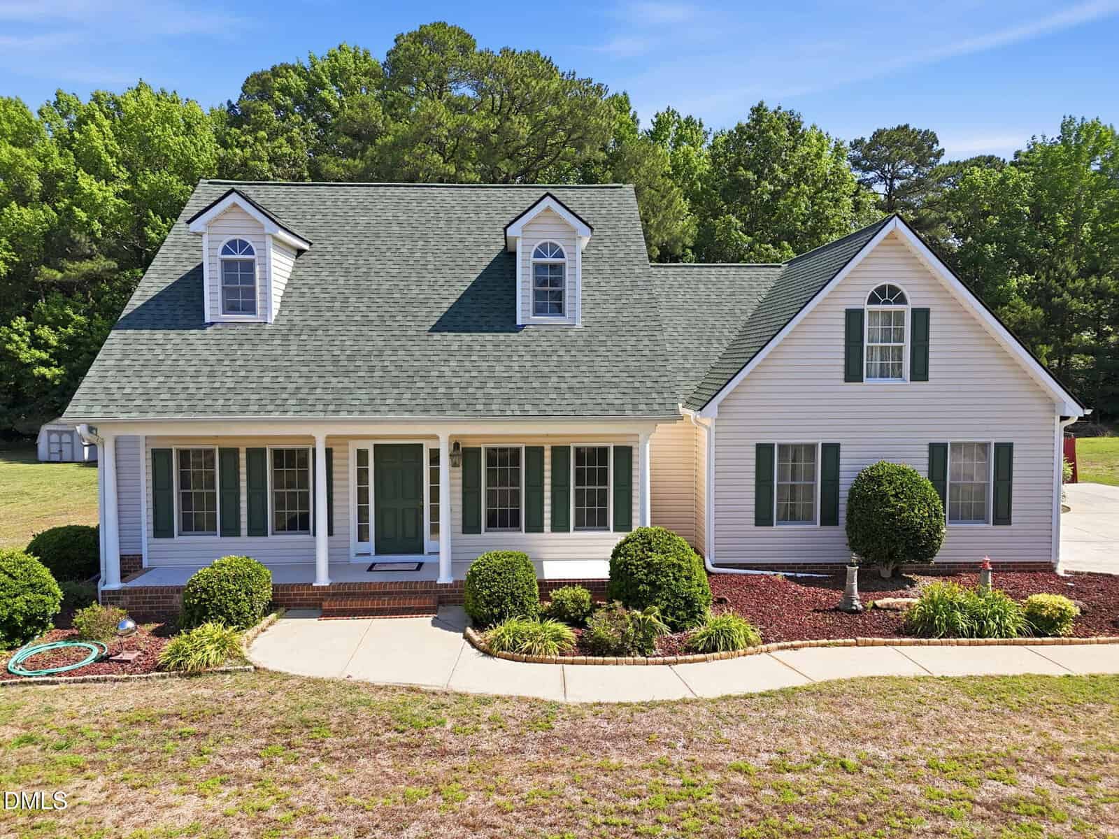 A white, two-story house at 120 Williamston Ridge Drive in Youngsville features green roof shingles, dormer windows, and a front porch. The yard has neatly trimmed bushes, a curved walkway, and is surrounded by trees under a clear blue sky.