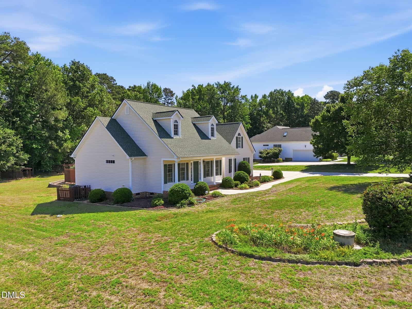 A white house with a green roof, front porch, and dormer windows sits on a spacious, grassy yard with bushes and a stone-lined flower bed. Another house and trees are visible in the background under a blue sky.