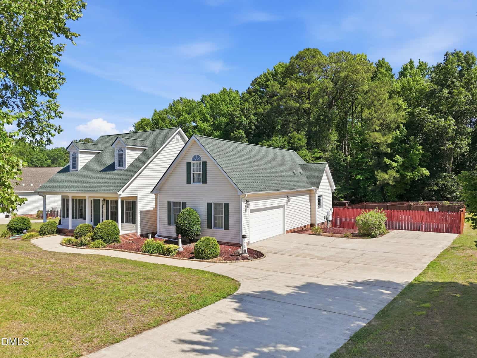 A white, single-story house with a green roof, attached garage, manicured shrubs, and a wide driveway. A red wooden fence encloses part of the backyard, bordered by tall trees under a clear blue sky.