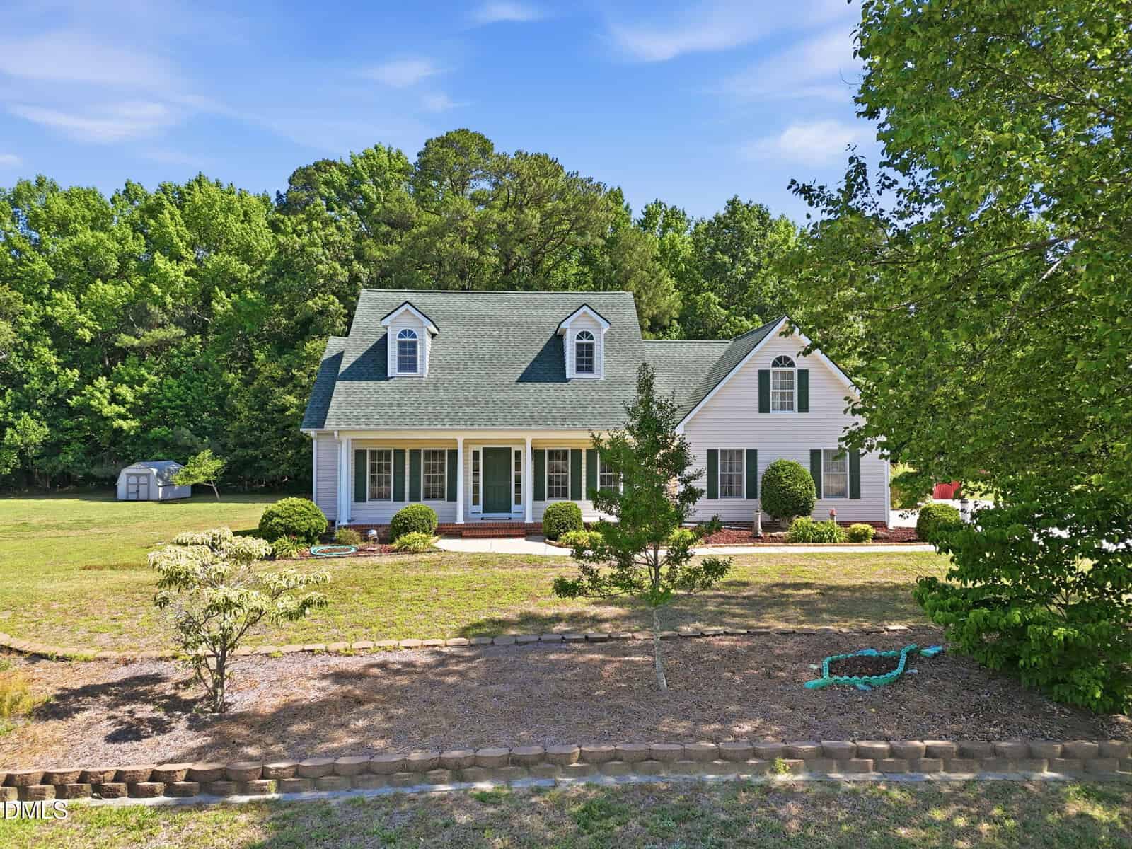 A white, two-story house with green roof sits on a spacious lot with a front porch, trimmed bushes, and surrounded by trees under a blue sky. A small storage shed is visible in the background.