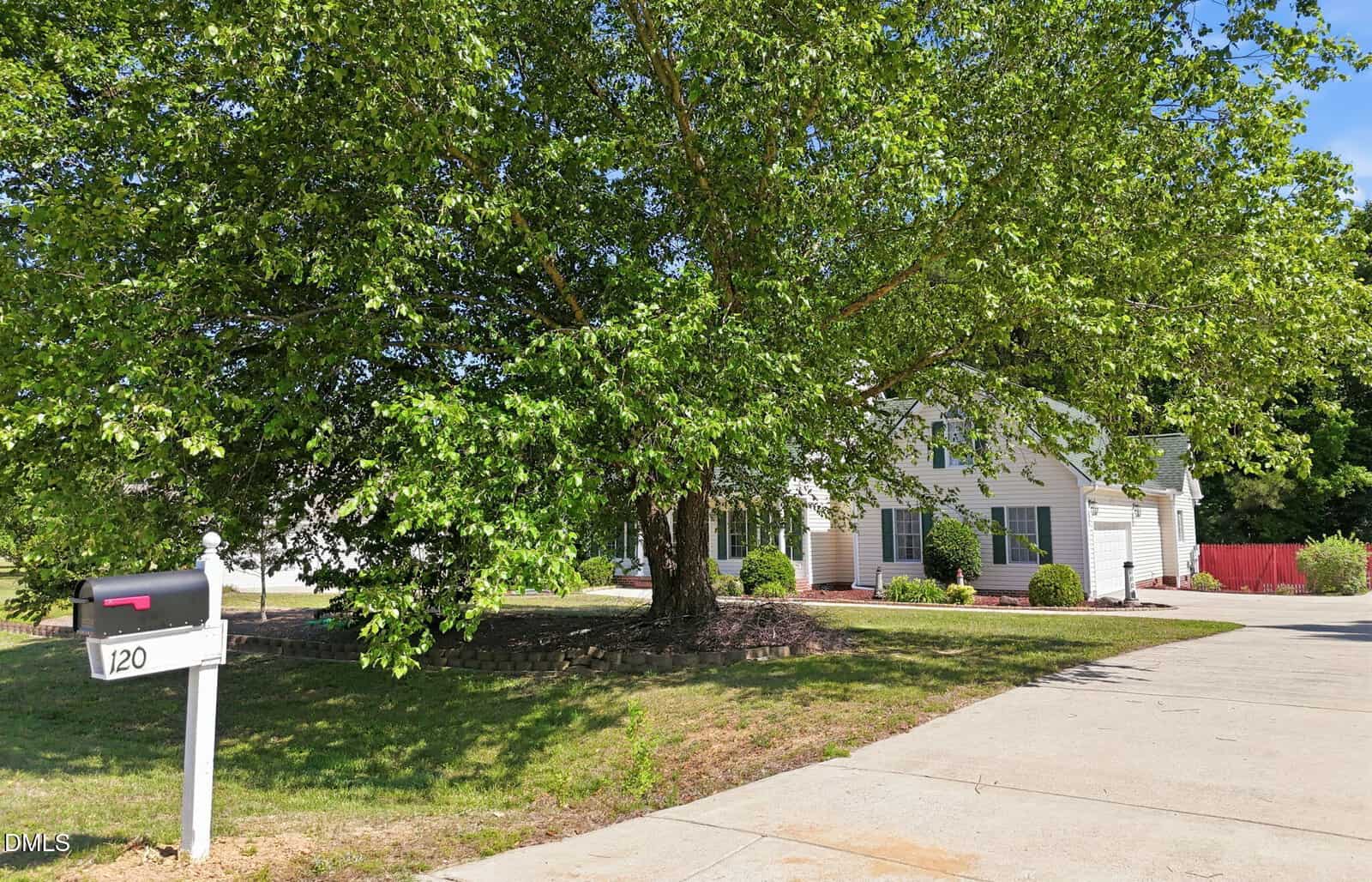 A white house with green shutters is partially shaded by a large leafy tree in the front yard. A black mailbox marked "120" stands near the driveway, and trimmed bushes line the house. It's a sunny day.
