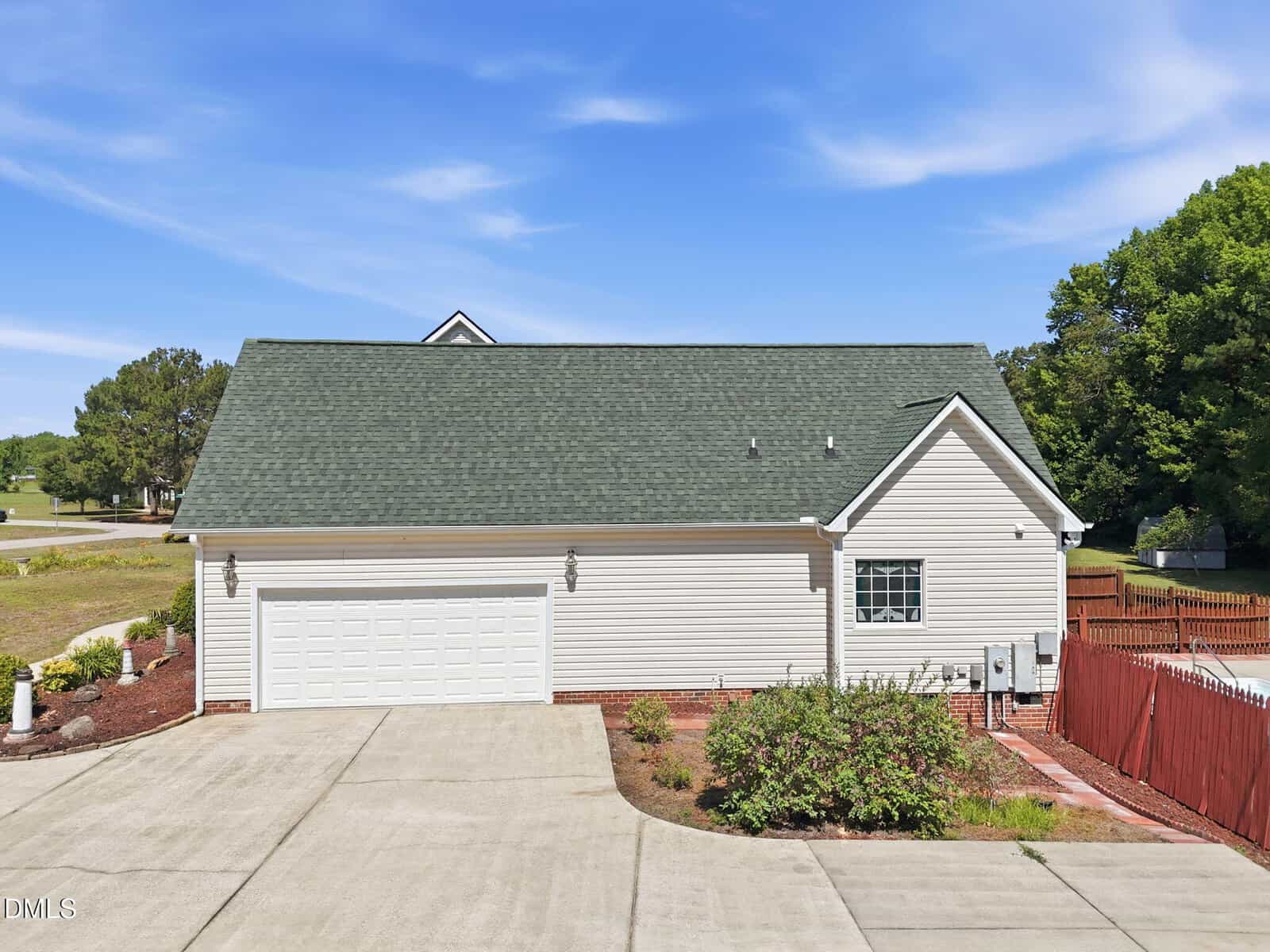 A beige house with a green roof, double garage doors, concrete driveway, small shrubs, and a red fence, set against a blue sky and surrounded by trees.
