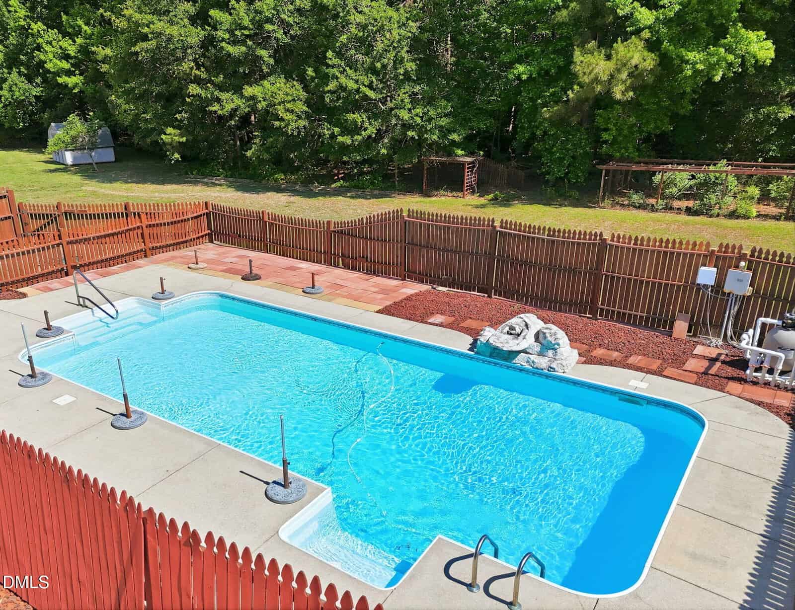 A rectangular in-ground swimming pool with clear blue water is surrounded by a wooden fence, a concrete deck, and trees in the background on a sunny day.