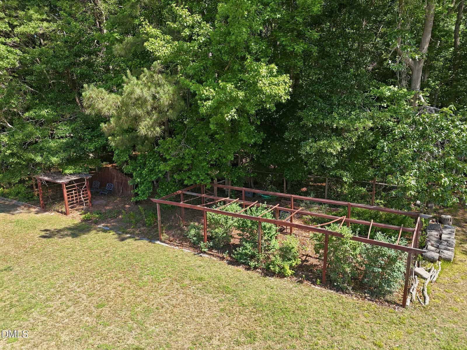 A fenced garden with raised beds and trellises sits beside a shaded seating area under trees, bordered by a grassy lawn and dense green forest in the background.