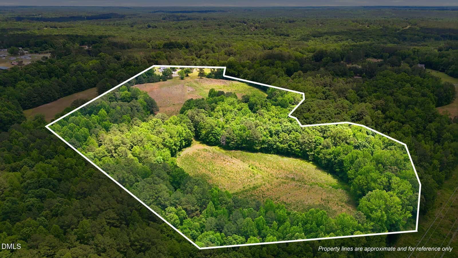 Aerial view of a large, wooded property with open clearings, outlined in white to indicate approximate boundaries, surrounded by dense forest and distant hills under a cloudy sky.