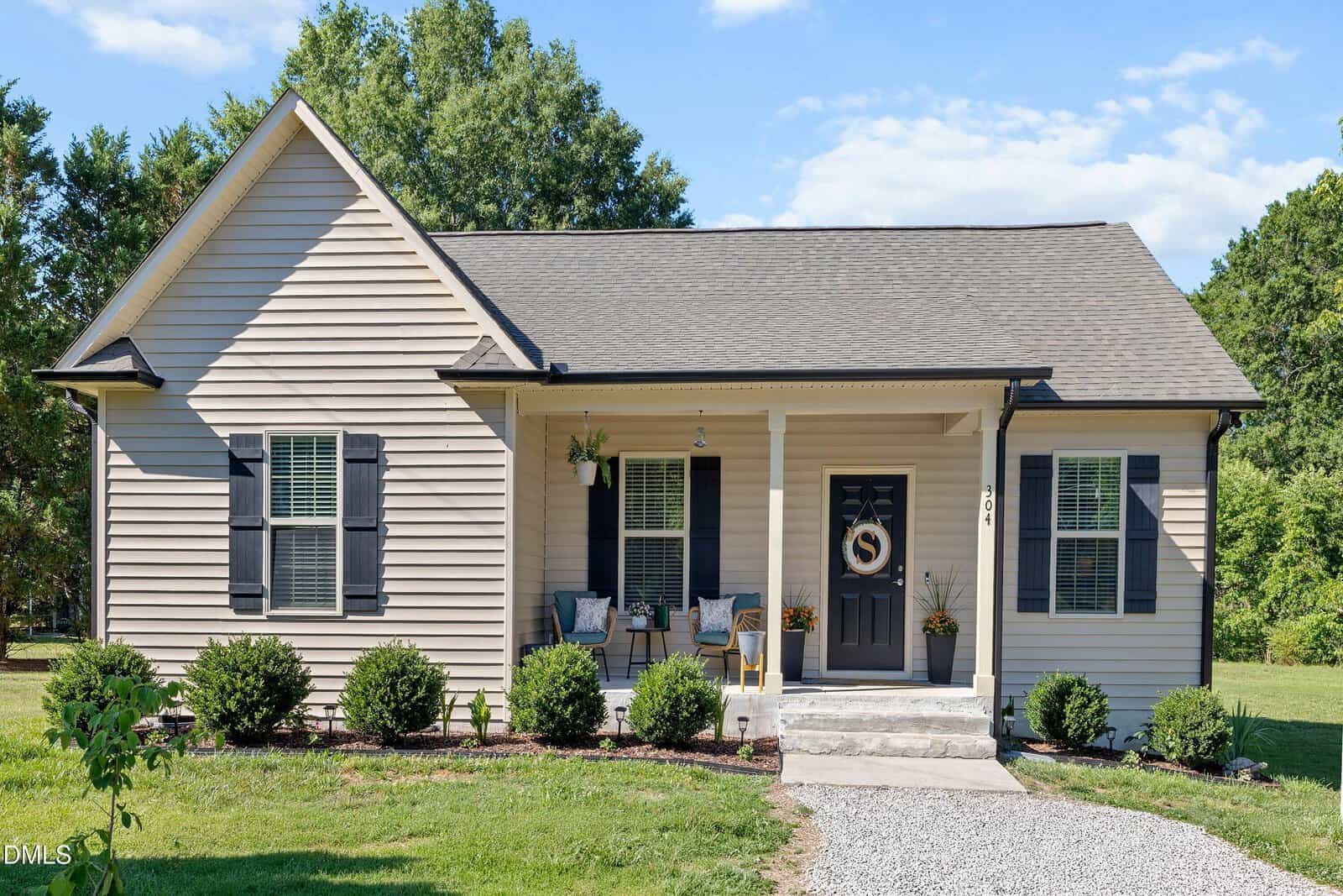 Nestled at 304 E Mason Street in Franklinton, this small beige house features dark shutters, a welcoming front porch with two chairs and potted plants, tidy shrubs, a gravel path to the black door, all surrounded by lush grass and trees.