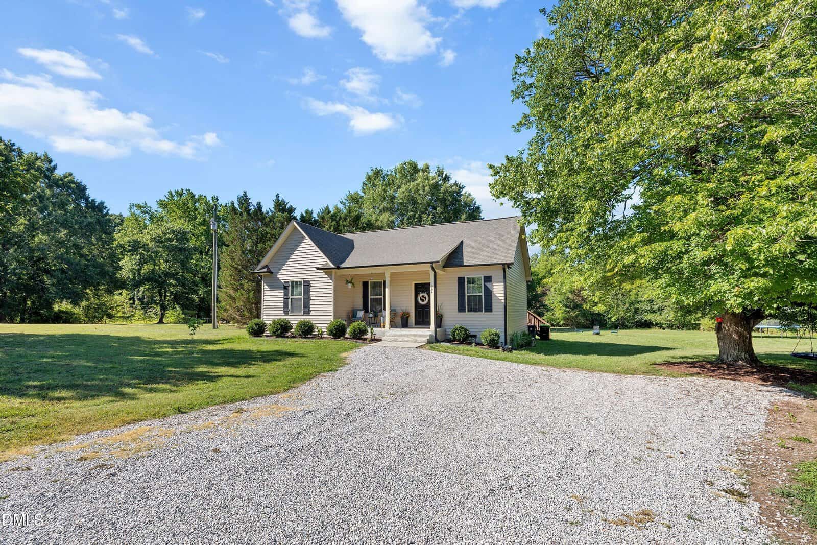 A small beige house with a front porch, gable roof, and wreath on the door sits on a spacious lot with a gravel driveway, surrounded by green grass and large trees under a blue sky with scattered clouds.