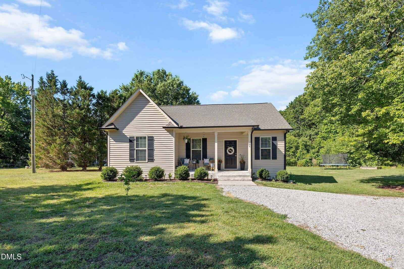 Single-story beige house with a covered front porch, gray roof, and white trim, surrounded by green grass, trees, and a gravel driveway under a blue sky with scattered clouds.