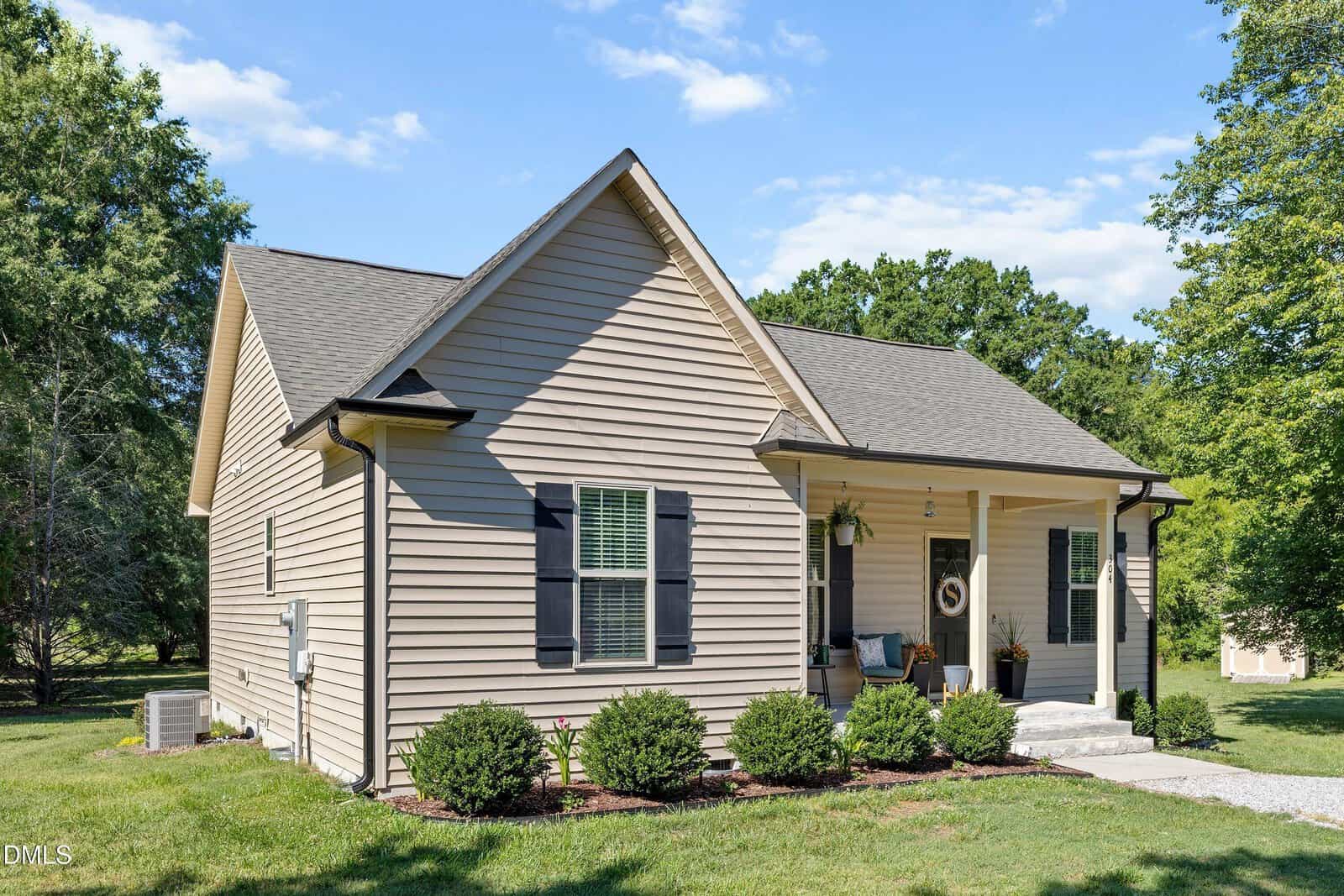 A small beige house with black shutters and a gabled roof, featuring a covered front porch with hanging plants and a wreath on the door, surrounded by green grass, shrubs, and trees under a blue sky.