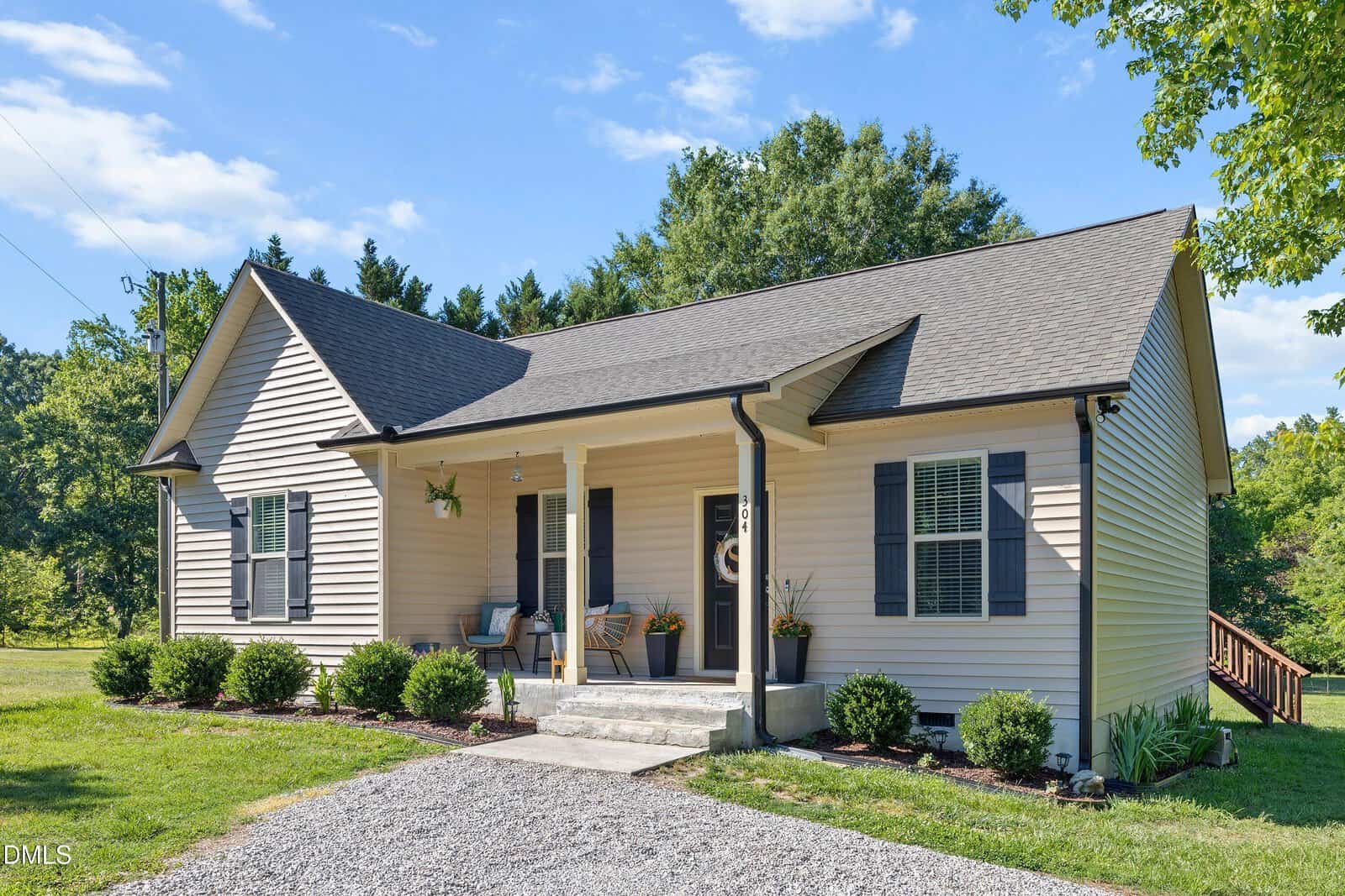 Single-story beige house with dark shutters and a covered front porch. There are potted plants and chairs on the porch, bushes along the front, and a gravel driveway. Trees and grass surround the home under a blue sky.