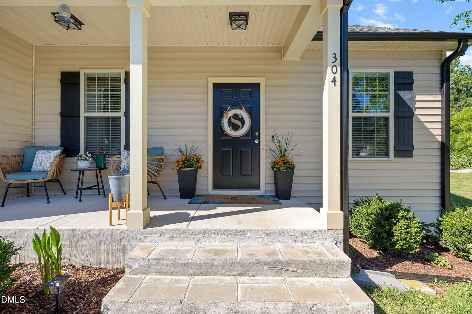 Beige house with black shutters, a black door displaying a gold "S" wreath, two potted plants, and a welcome mat. Porch with wicker chairs, pillows, and small table. Stone steps lead to a landscaped front yard.