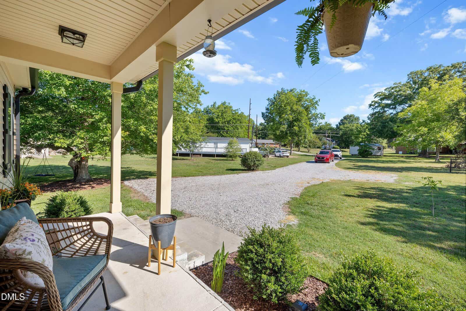 View from a covered porch showing a wicker bench, potted plants, a gravel driveway, a red car, a white trailer, green lawn, trees, and a clear blue sky. Shadows from the porch roof fall across the concrete steps.
