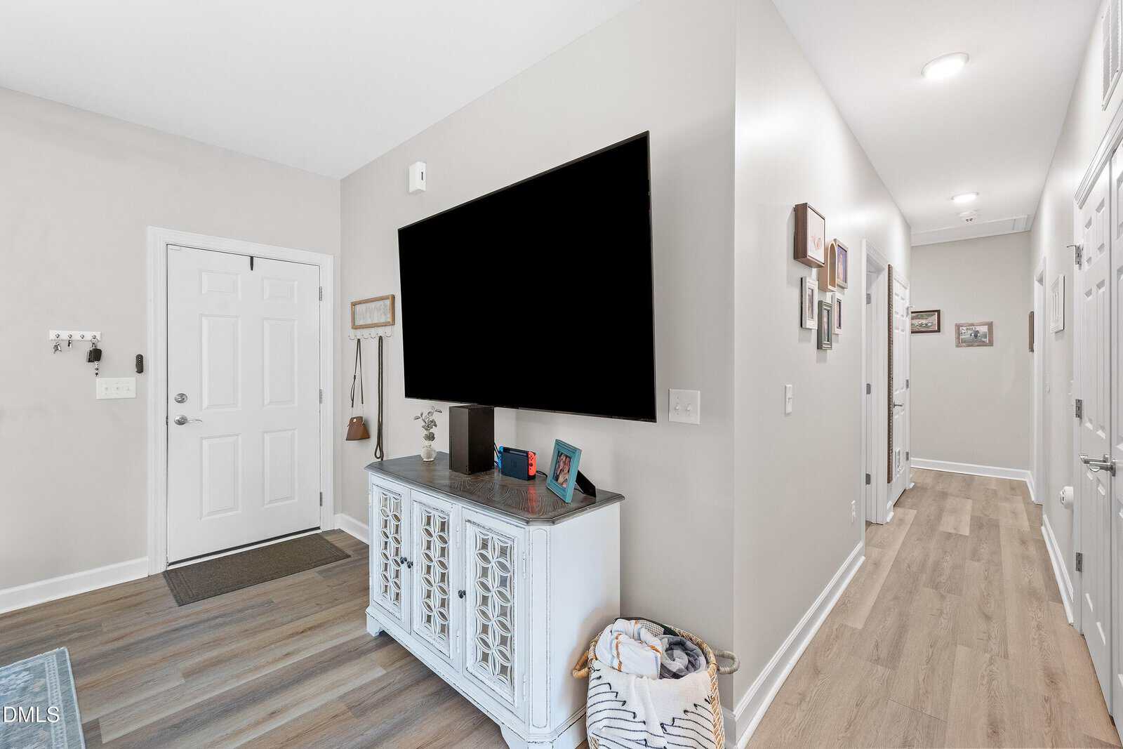 A hallway with light wood floors, neutral walls, a closed white door, a large flat-screen TV on a white console table, framed pictures on the wall, and a woven basket on the floor.