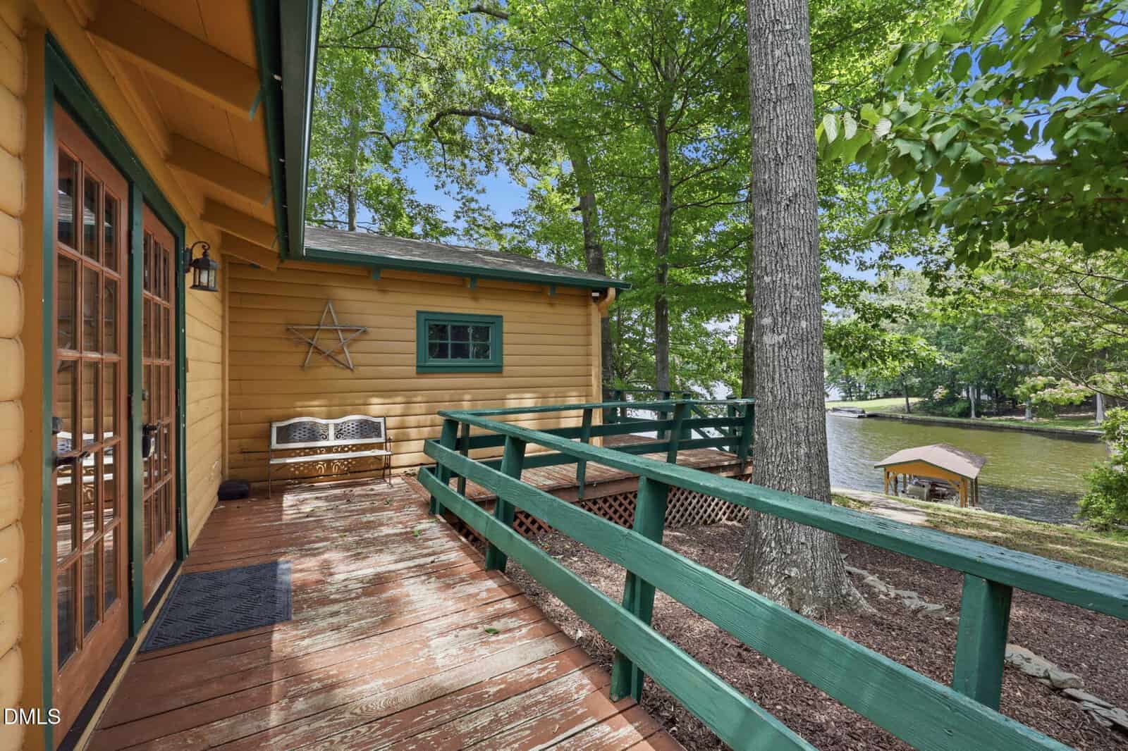 A wooden cabin with a deck and green railings overlooks a lakeside view surrounded by trees. A wooden bench sits on the deck, and a dock with a boat is visible on the lake below.