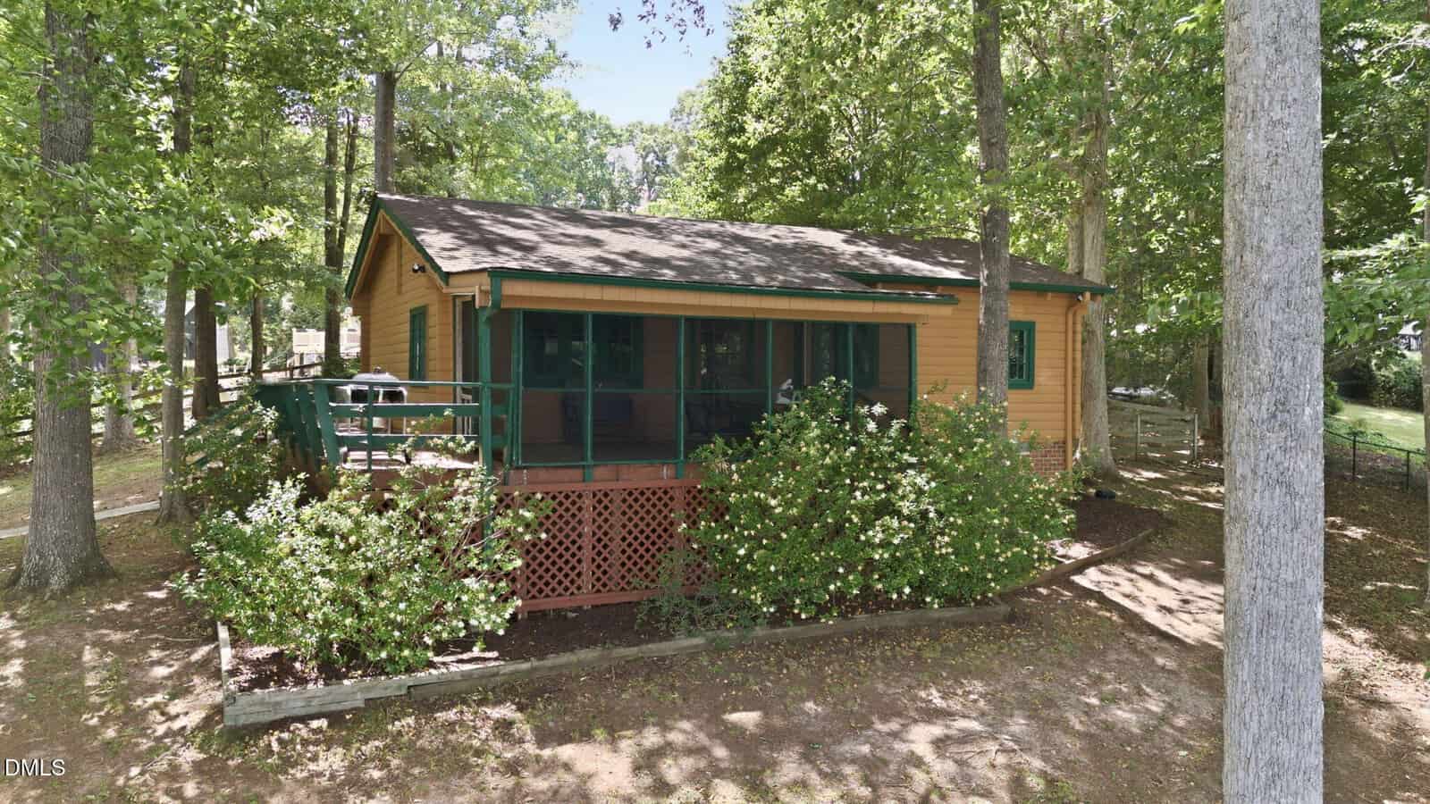 A small wooden cabin with a screened porch and green railings is surrounded by trees and bushes. Sunlight filters through the leaves, casting dappled shadows on the ground.