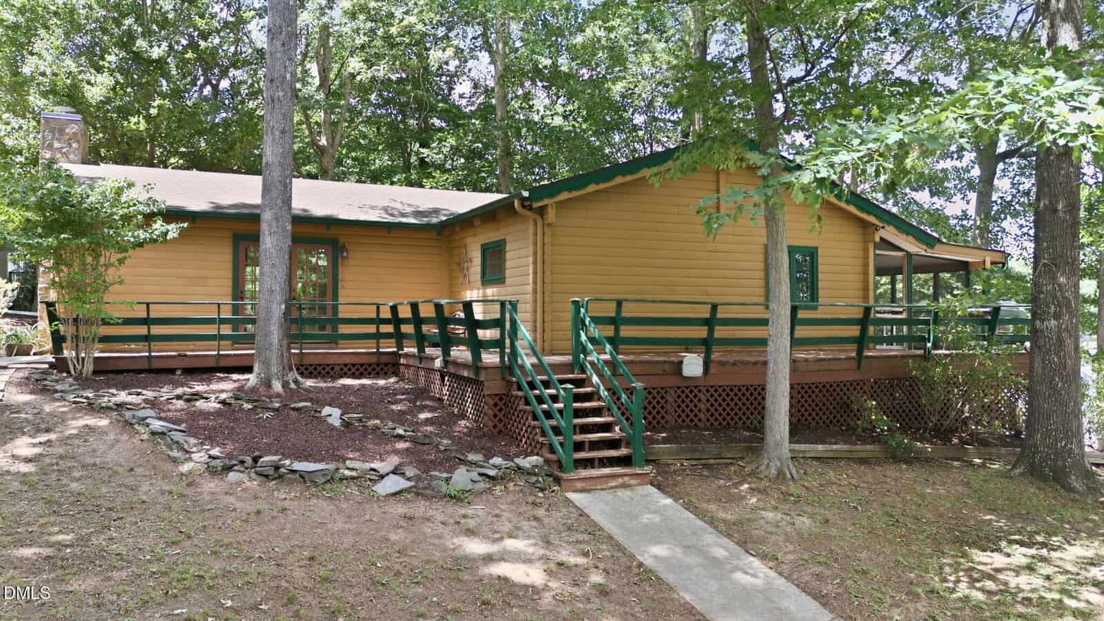 A wooden cabin with a green trim and large wraparound porch sits among tall trees. A ramp and stairs lead up to the porch, and the ground is covered with rocks and mulch. Sunlight filters through the tree canopy.