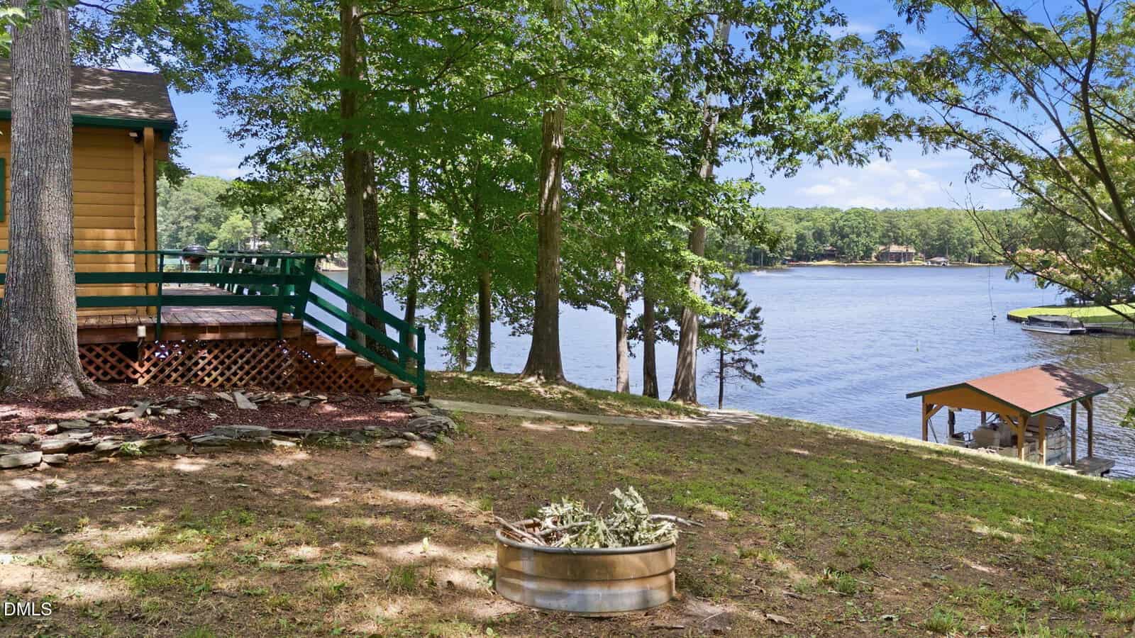 A lakeside cabin with a porch overlooks calm water, surrounded by tall trees. A wooden dock and covered boat lift are visible on the right, with a metal planter in the grassy yard.